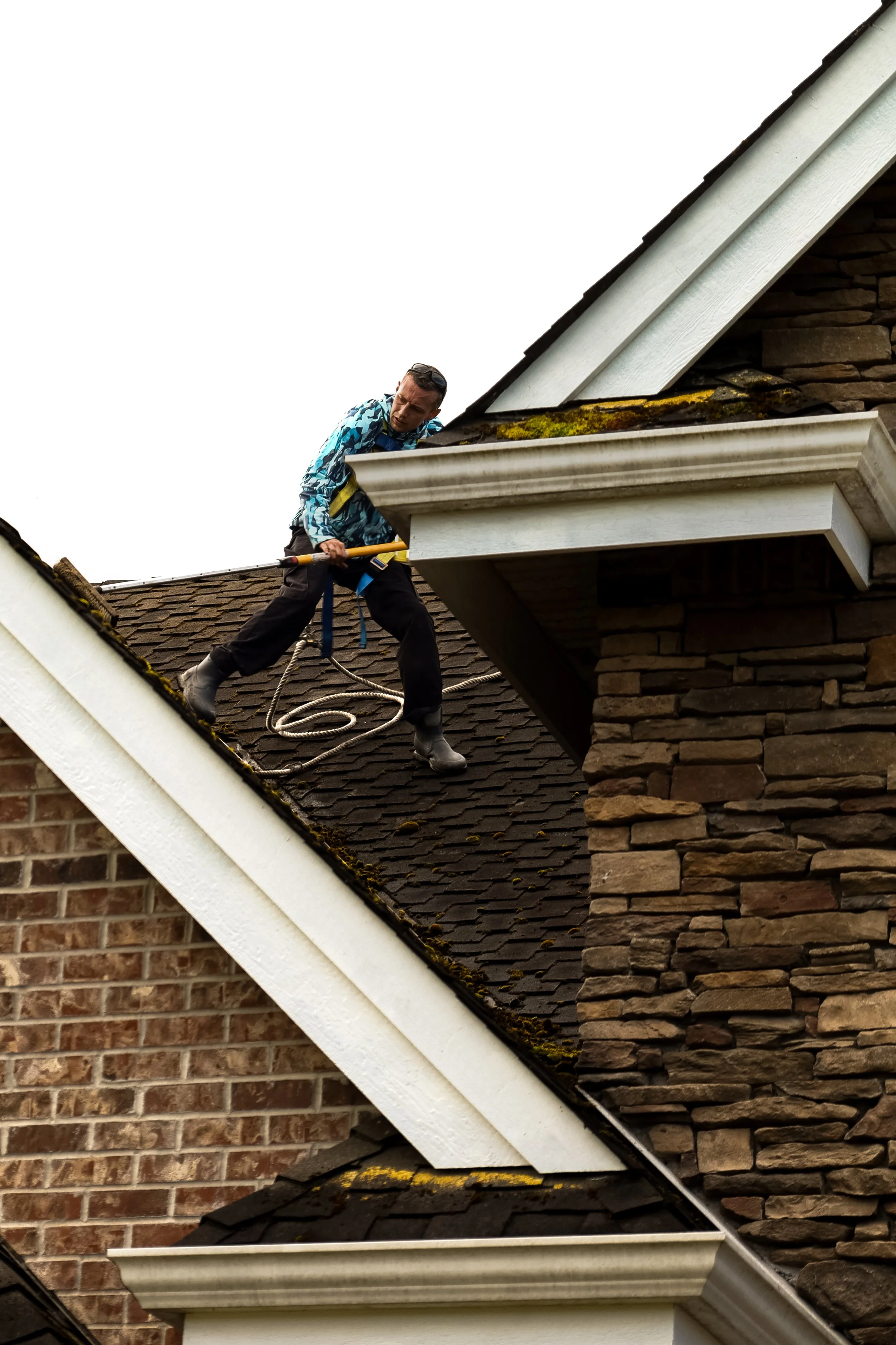 A man wearing a blue patterned jacket, black pants, and white boots is on a roof, using a tool and rope while inspecting or fixing the roof. The house has brown shingles, a chimney made of stone, white trim, and moss on the roof.
