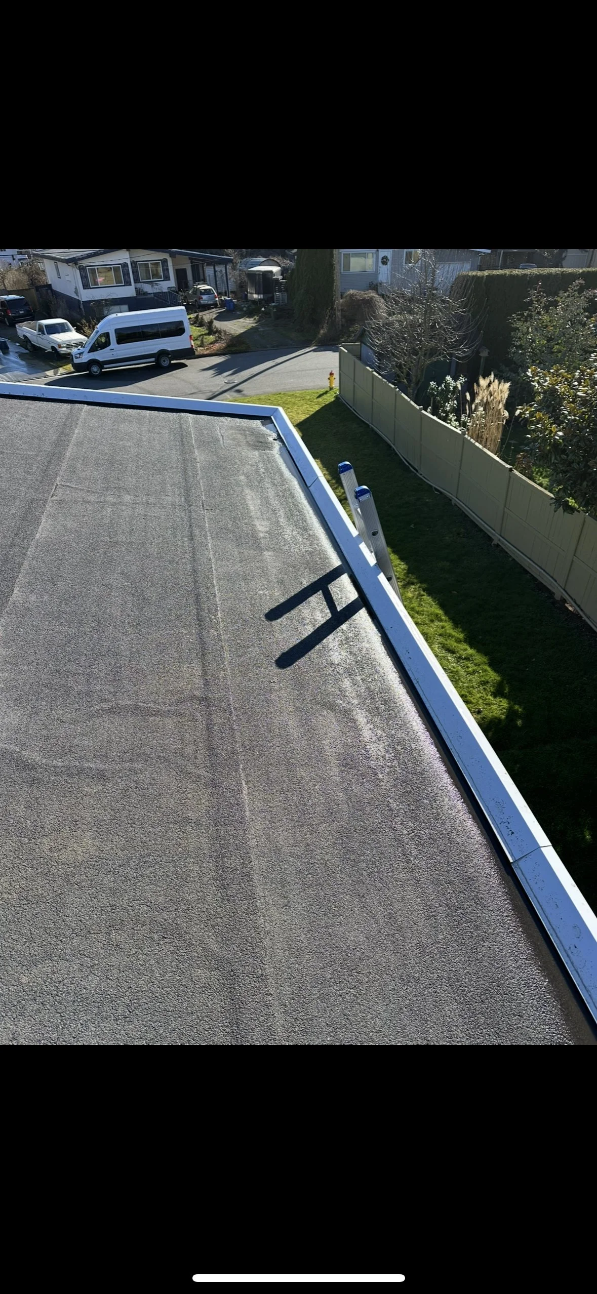A rooftop with a textured surface and a ladder leaning against the edge, casting a shadow on the roof, with neighboring houses, vehicles, and a street visible in the background.