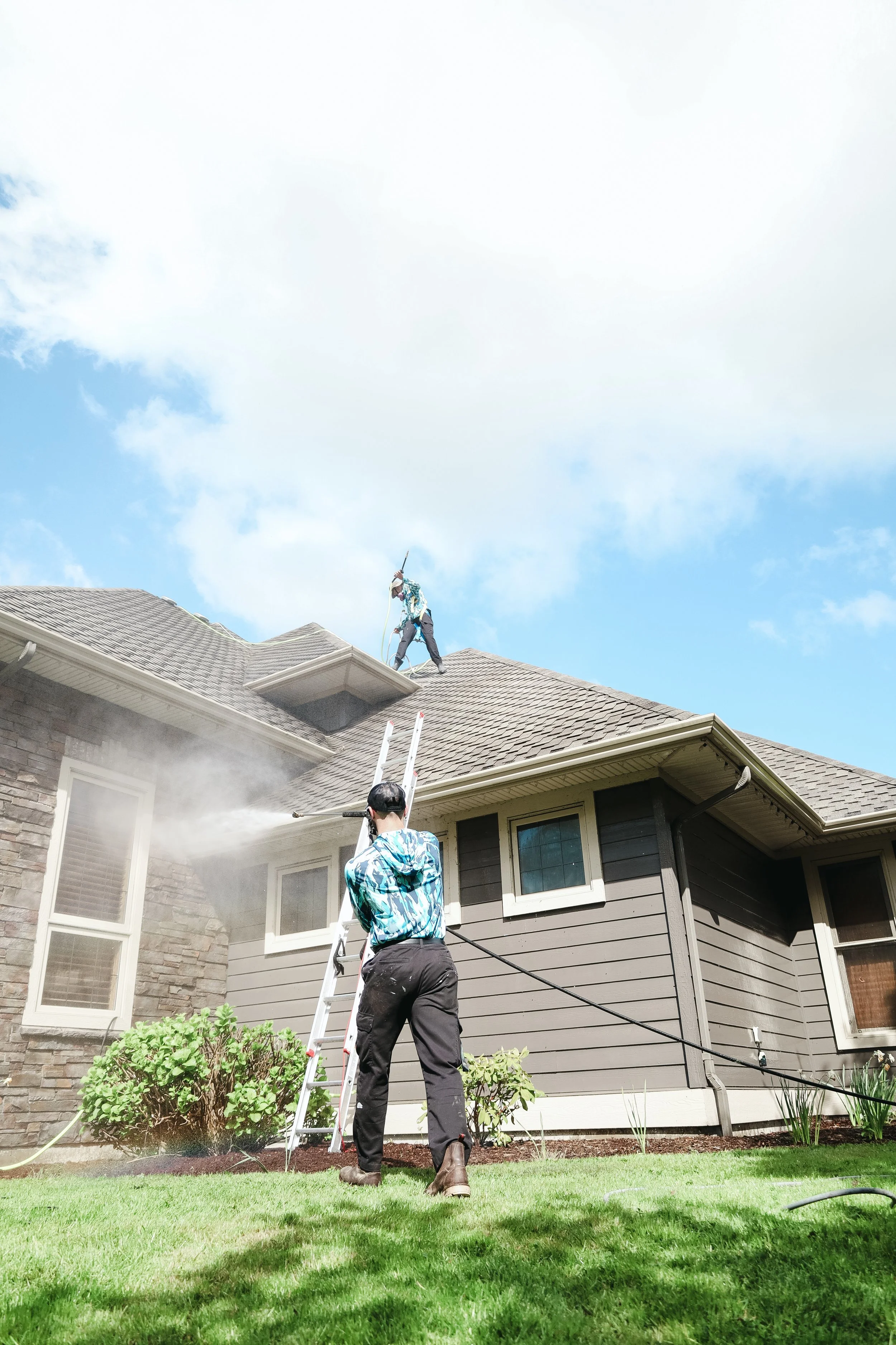 Two people cleaning the roof of a house with tools, one on a ladder and the other on the ground, while a third person stands nearby holding a ladder, with a house and a lawn in the background.