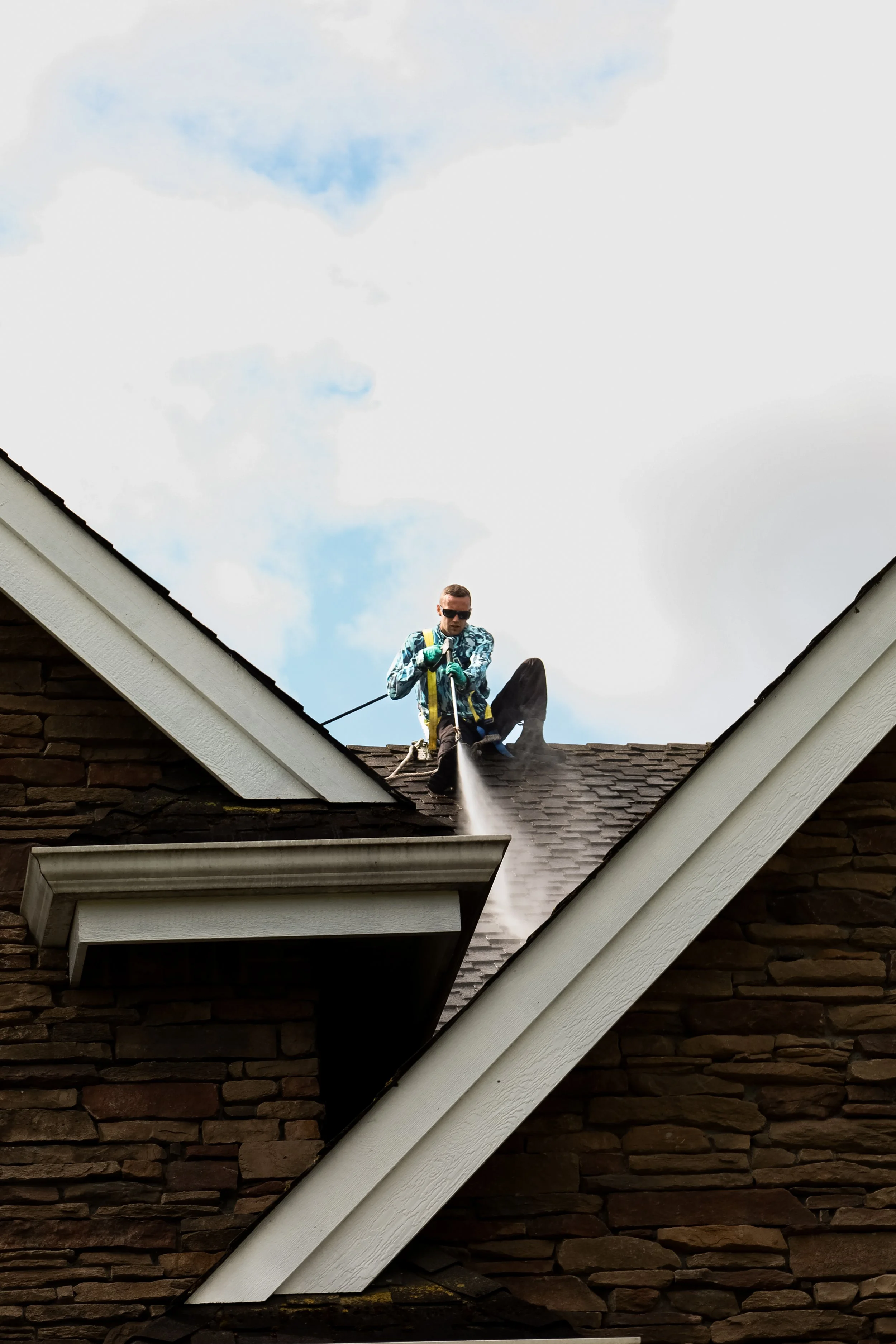 Man wearing sunglasses, a patterned jacket, and gloves on a roof, cleaning or inspecting the shingles with a hose or tool during daytime.