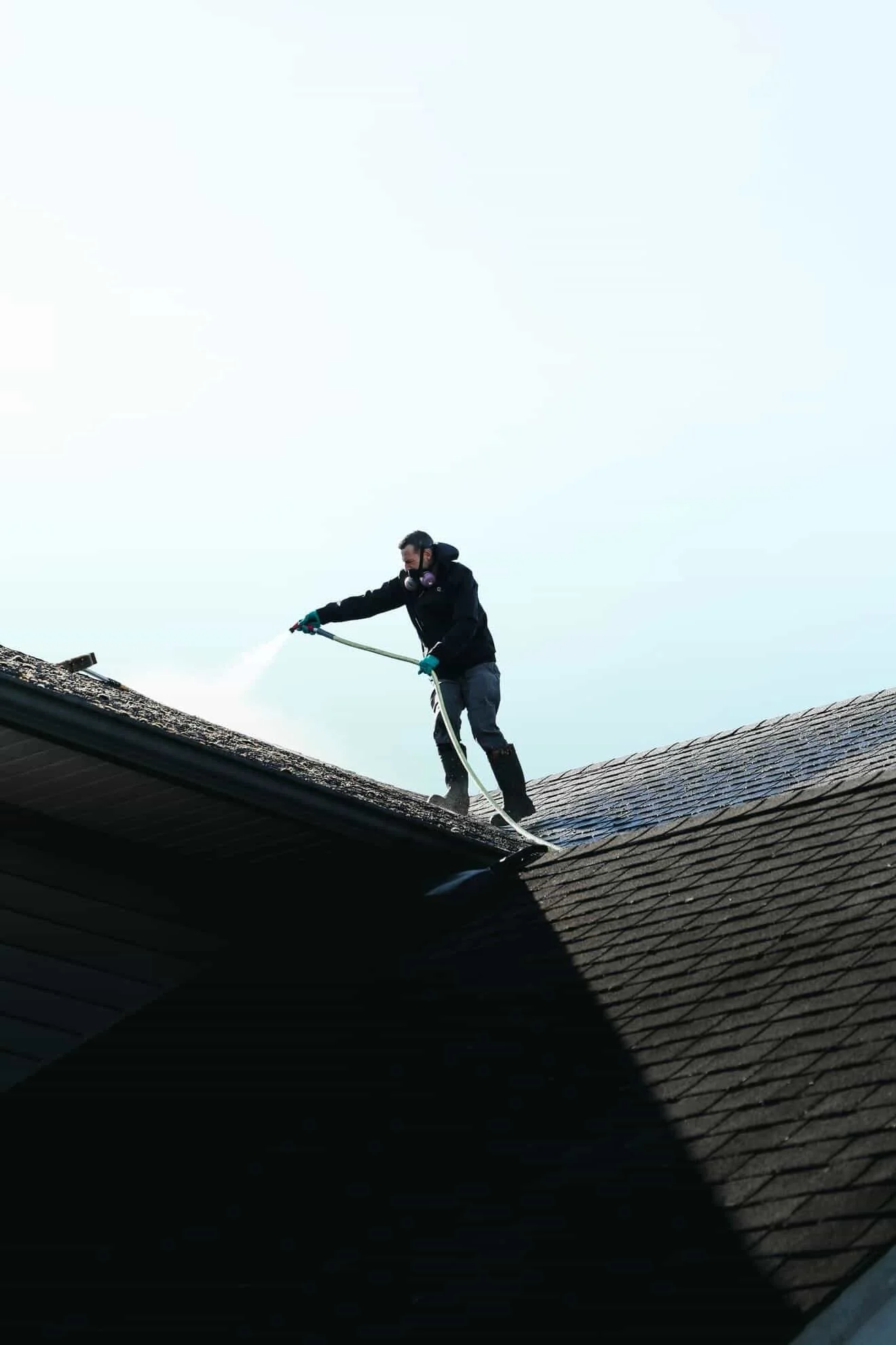 A man wearing a mask, gloves, and boots is cleaning a roof with a hose.
