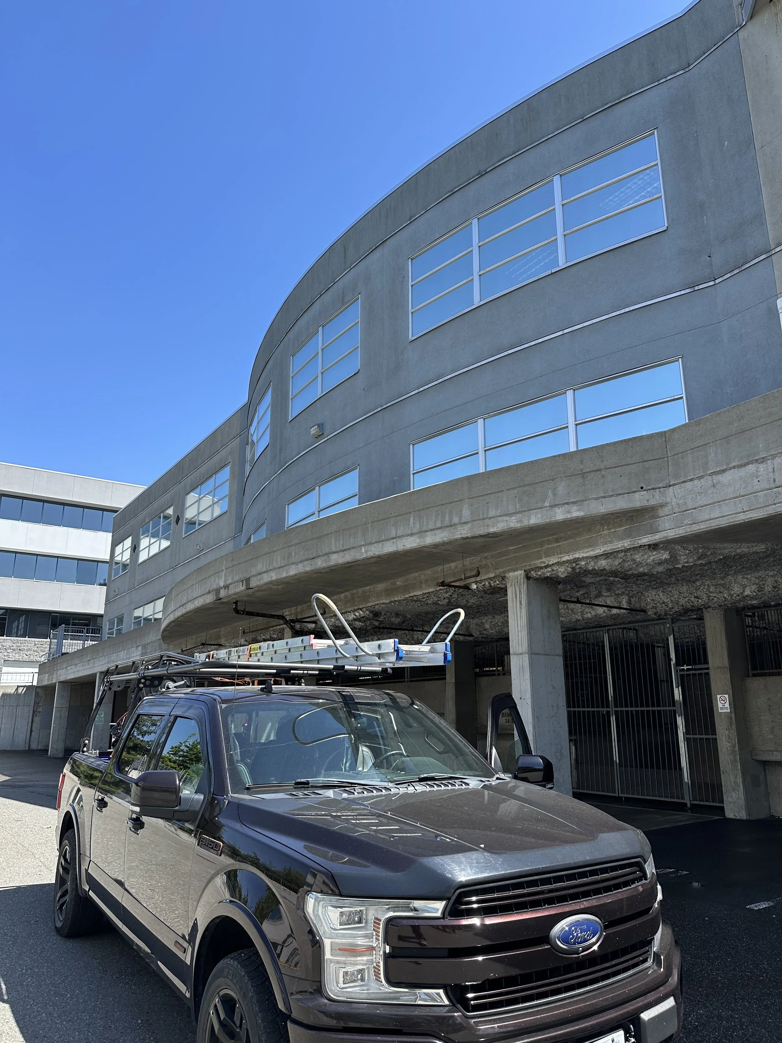 A gray Ford pickup truck parked in front of a modern, multi-story building with large glass windows and concrete exterior walls, under a clear blue sky.