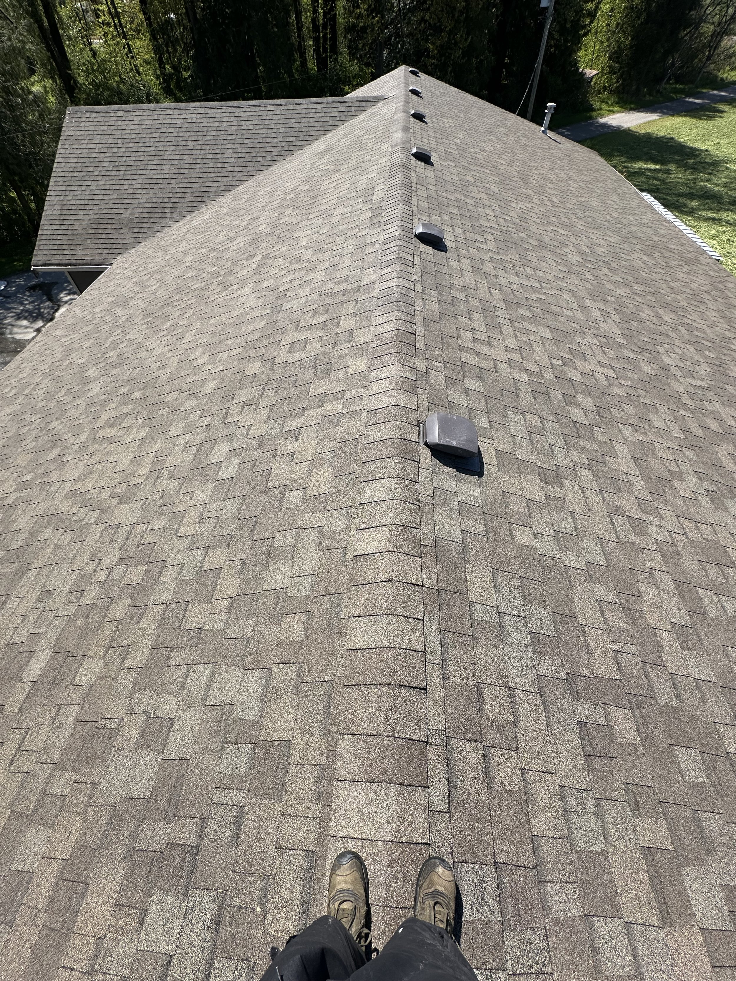 A person standing on a roof wearing hiking boots, with a view of the asphalt shingles, roof vents, and surrounding trees.