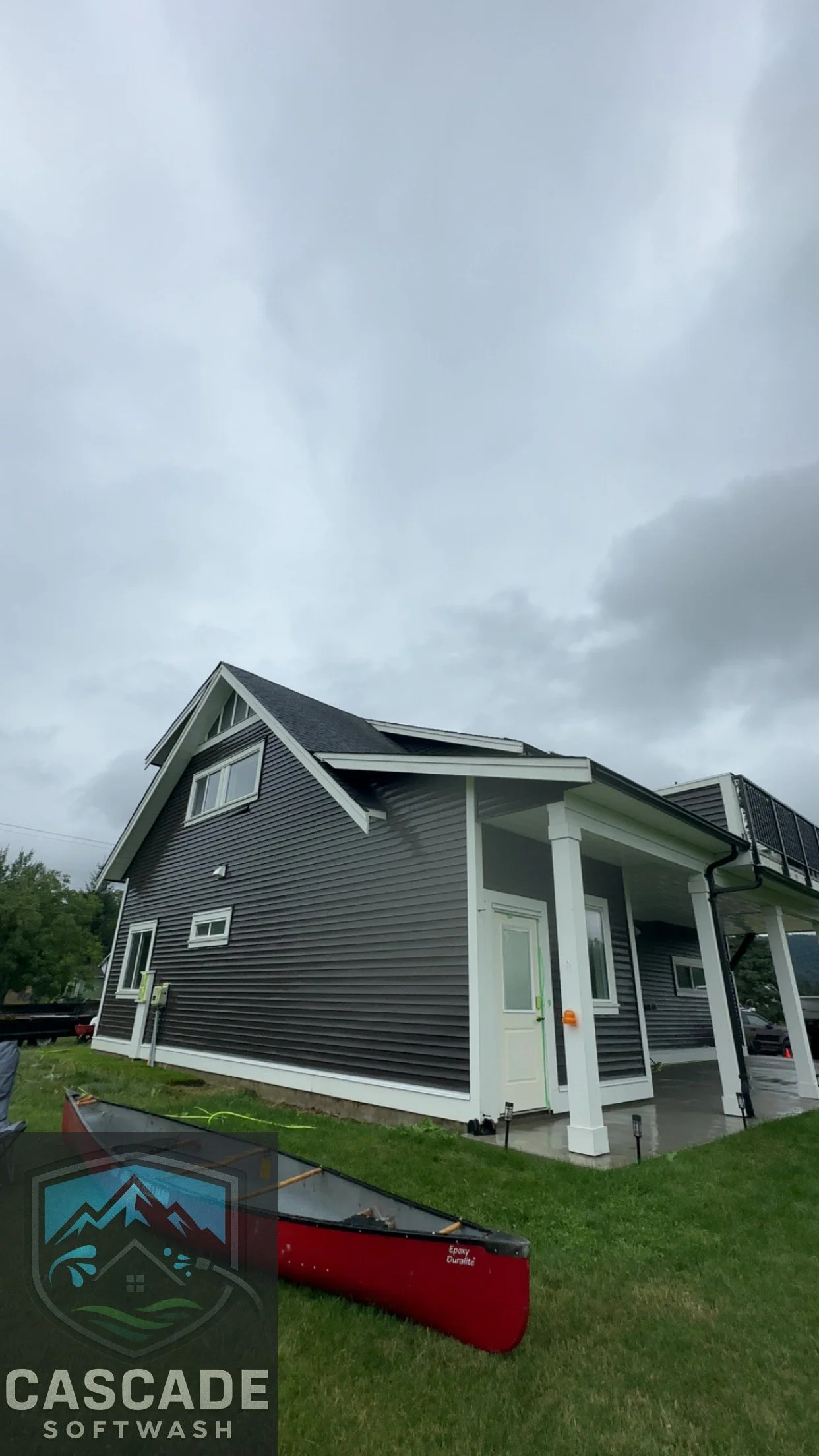 A two-story house with gray siding, white trim, and a gable roof under a cloudy sky. A white door and large windows are visible, with a small porch area supported by white columns. A red kayak is partially visible on the lawn, which is green and slig