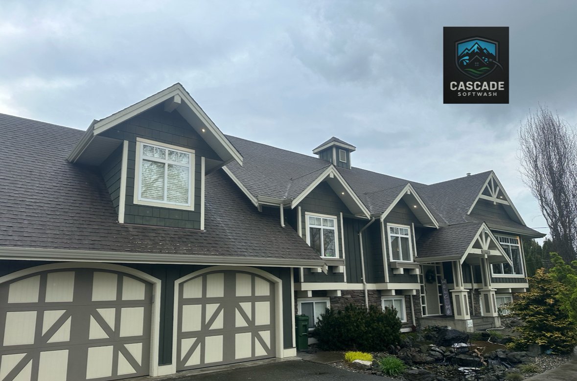 A large, multi-story house with green and beige exterior, multiple gabled roofs, and white-framed windows. There's a two-car garage with decorative doors, a driveway, and landscaped shrubs and rocks in the front yard. The sky is cloudy, and there's a