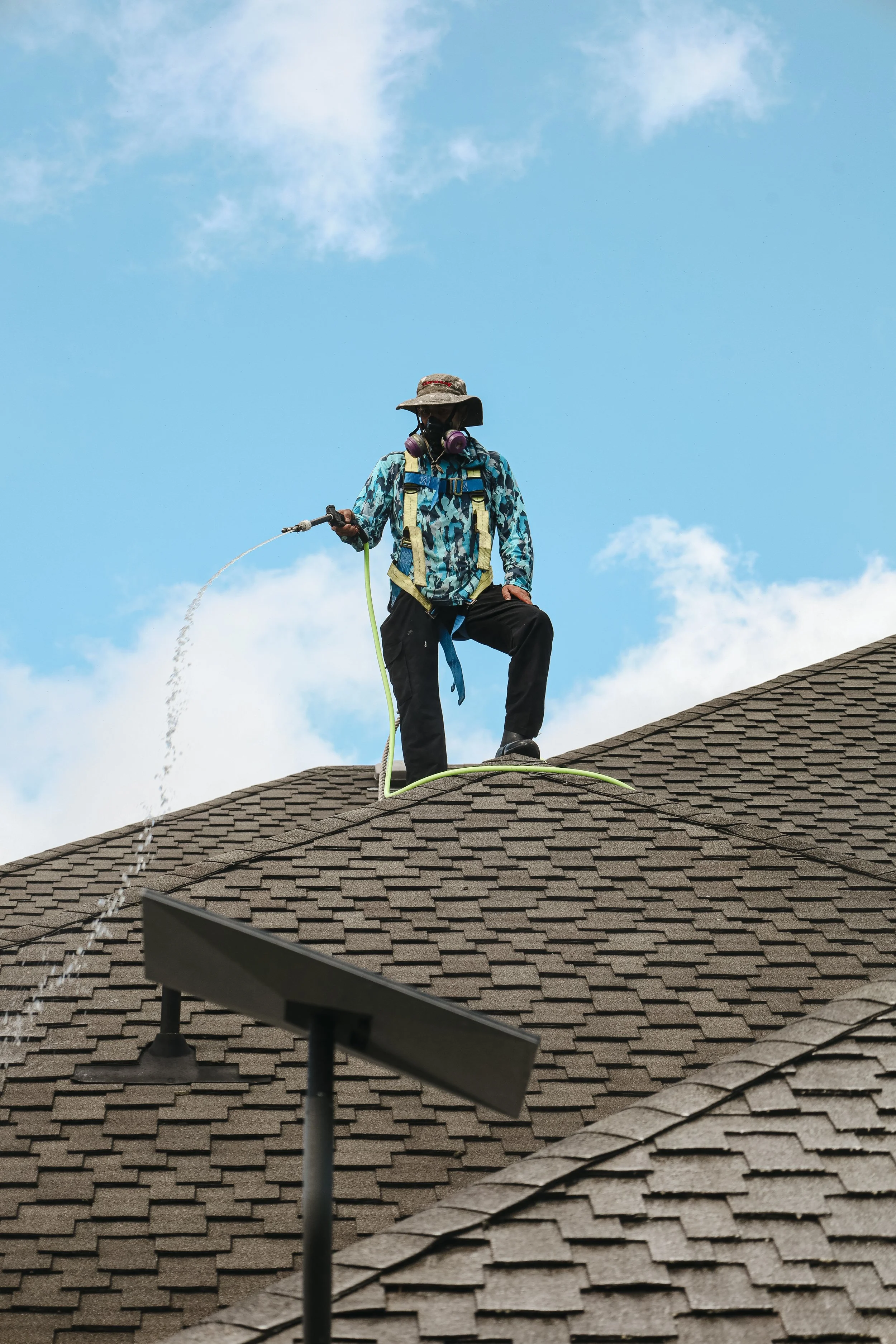 A person standing on a rooftop in a camouflage jacket, wearing a wide-brimmed hat, goggles, and a gas mask, using a hose to spray water on the roof against a blue sky with some clouds.