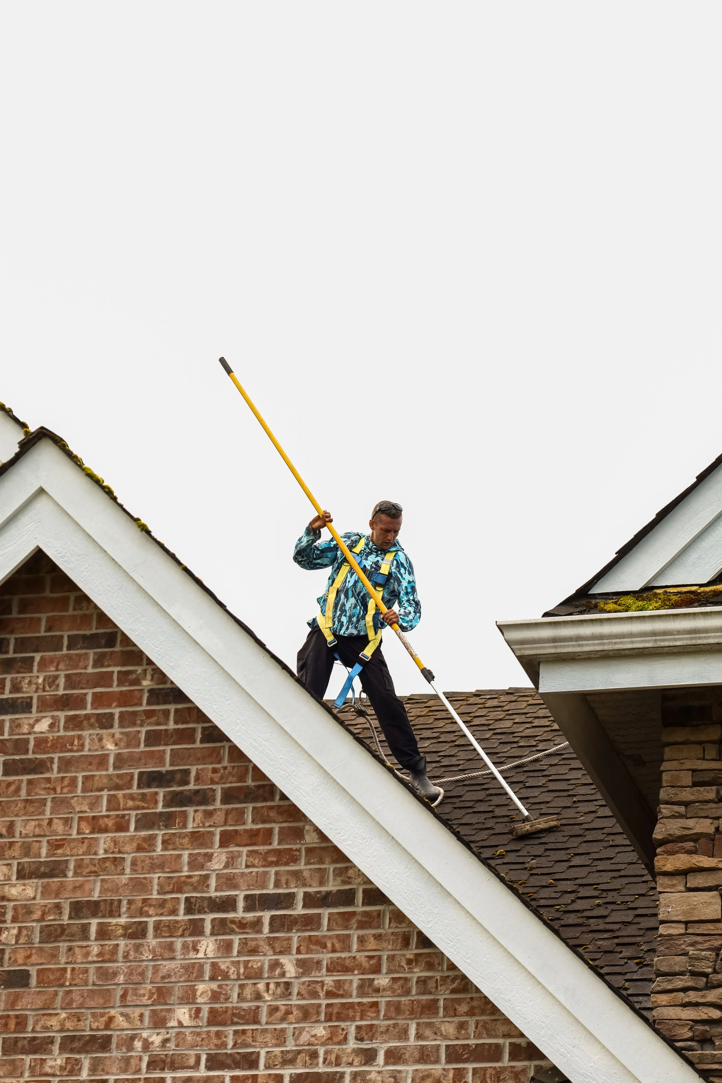 A person in a blue patterned jacket and black pants is working on the roof of a brick house, holding a yellow tool, wearing a safety harness, with a cloudy sky in the background.