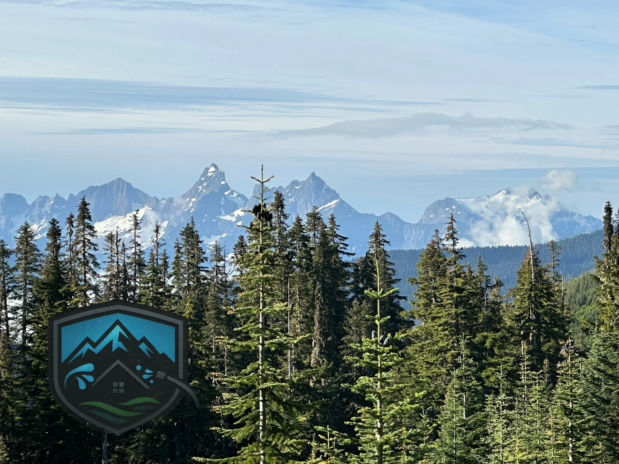 A scenic view of a dense forest of pine trees with snow-capped mountains in the background under a partly cloudy sky, featuring a logo with mountains, a house, and a water droplet.
