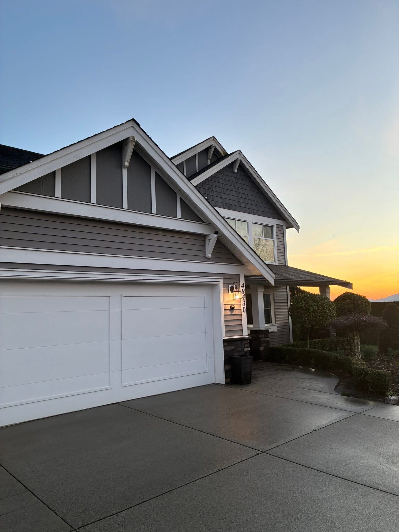 Exterior view of a modern two-story house at sunset, showing a white garage door, house number 48430, front door with a lit wall-mounted light, landscaped bushes, and a driveway.