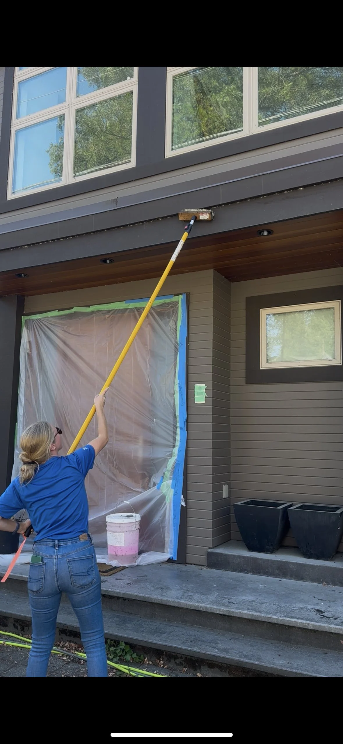 A woman in a blue shirt and jeans is using an extension pole to paint or clean the corner of a building's exterior near a window. The building has dark gray siding, a small window, and large planters on the porch.