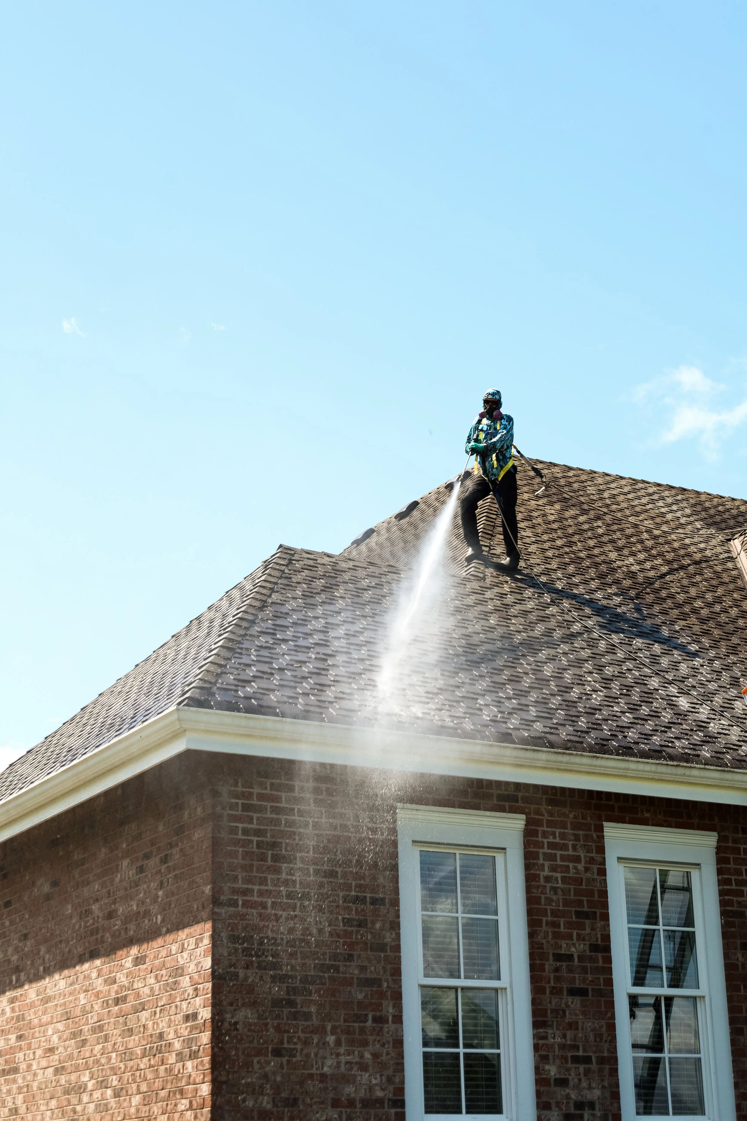 A person wearing work gear, including a safety mask and helmet, is pressure washing the roof of a brick house on a clear day.