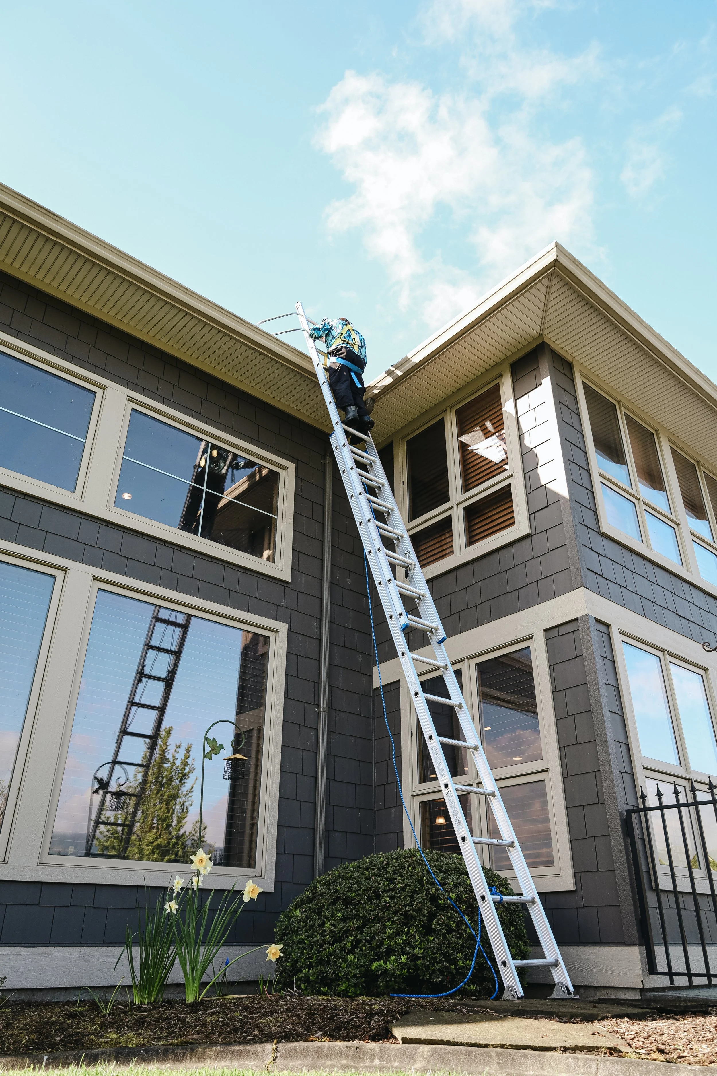 A person standing on a ladder cleaning or repairing the roof of a three-story house with dark gray siding. The house has large windows, and there are trees and flowers in the front yard under a partly cloudy sky.