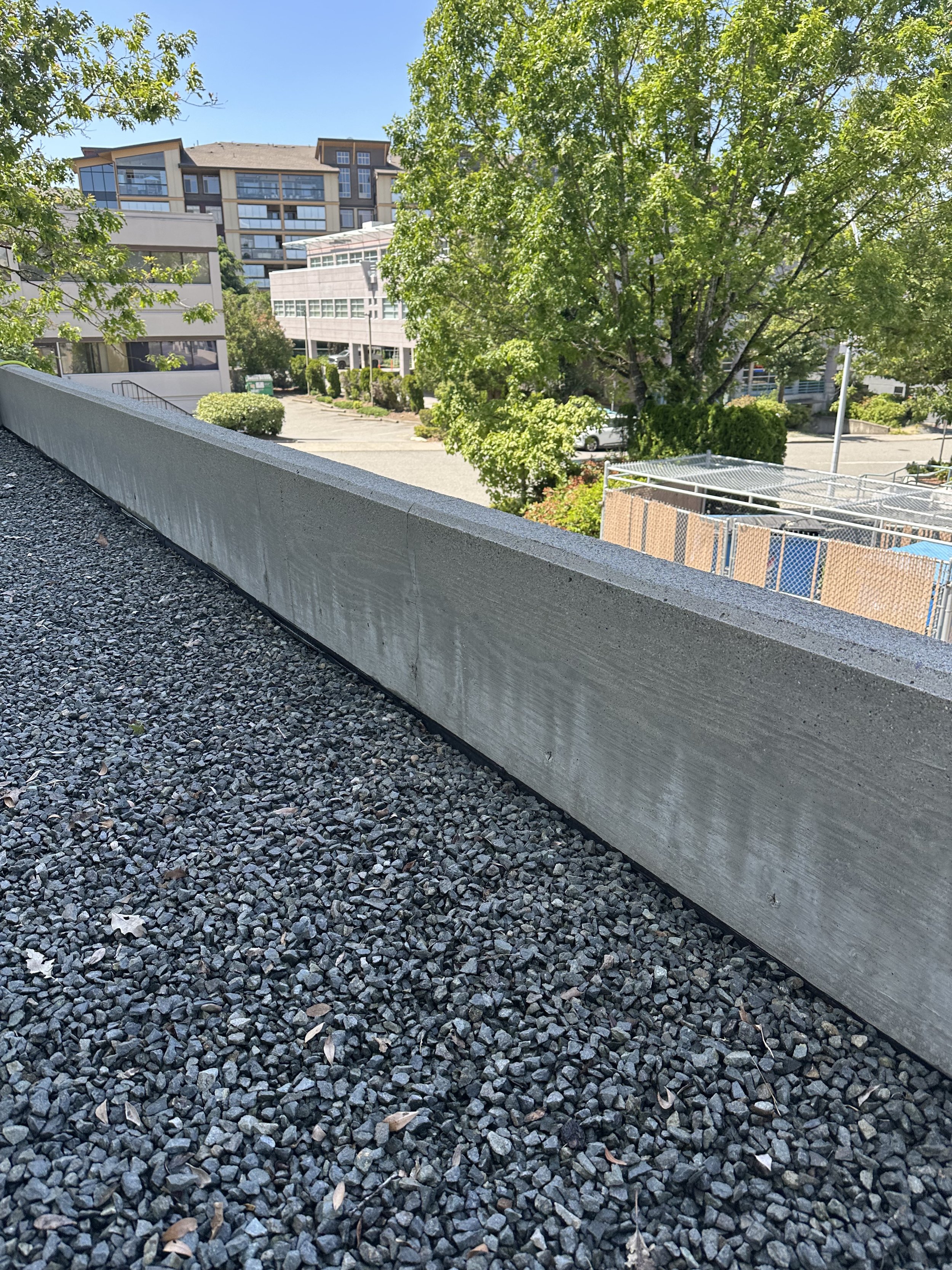 Cityscape view from a balcony with gravel foreground, concrete railing, trees, and modern buildings in the background under a clear blue sky.