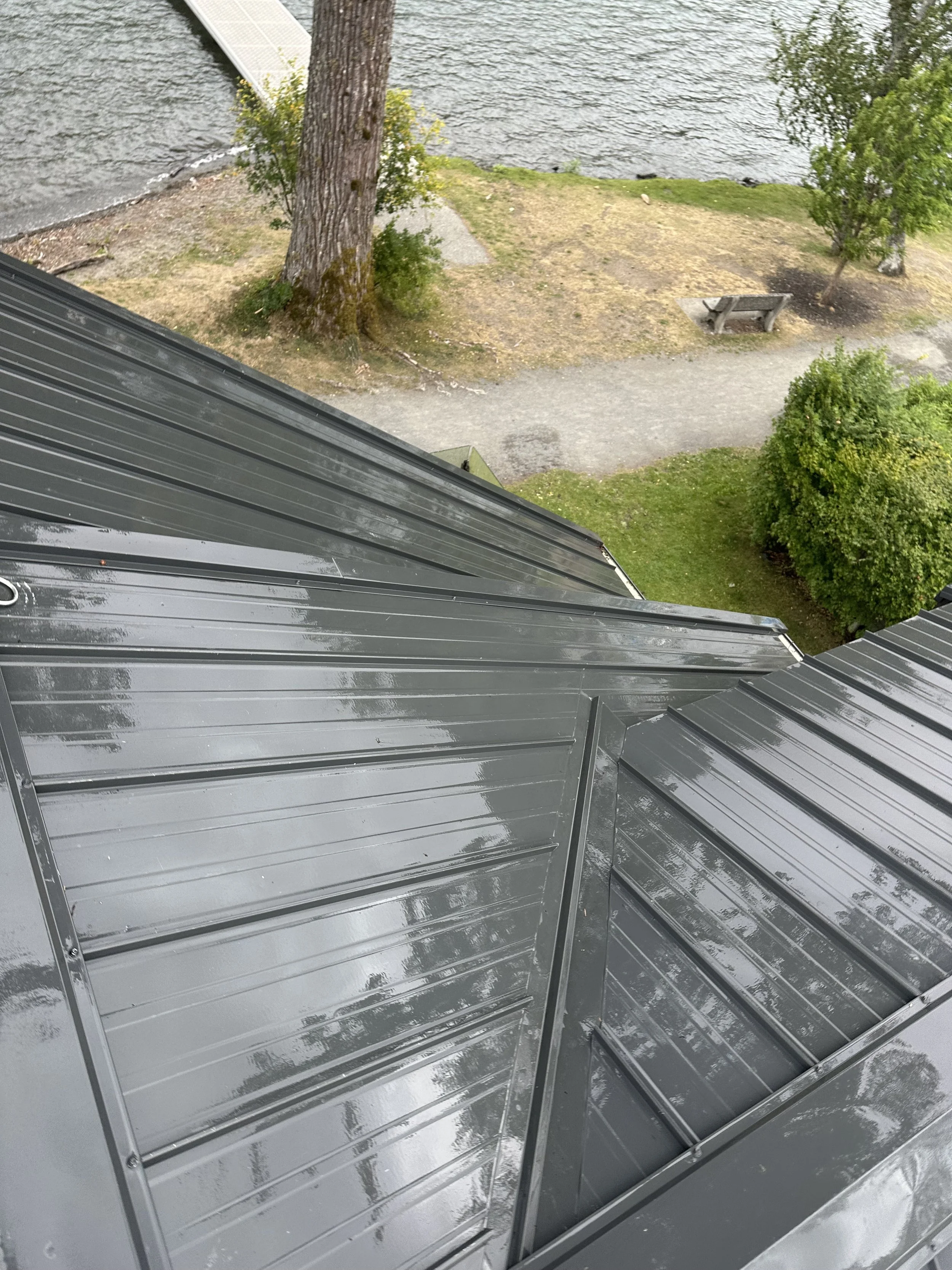 A photo of a sheet of metal roofing material with a shiny, reflective surface, beneath the edge of a roof, overlooking a park area with trees, a bench, and the water of a lake or river in the background.