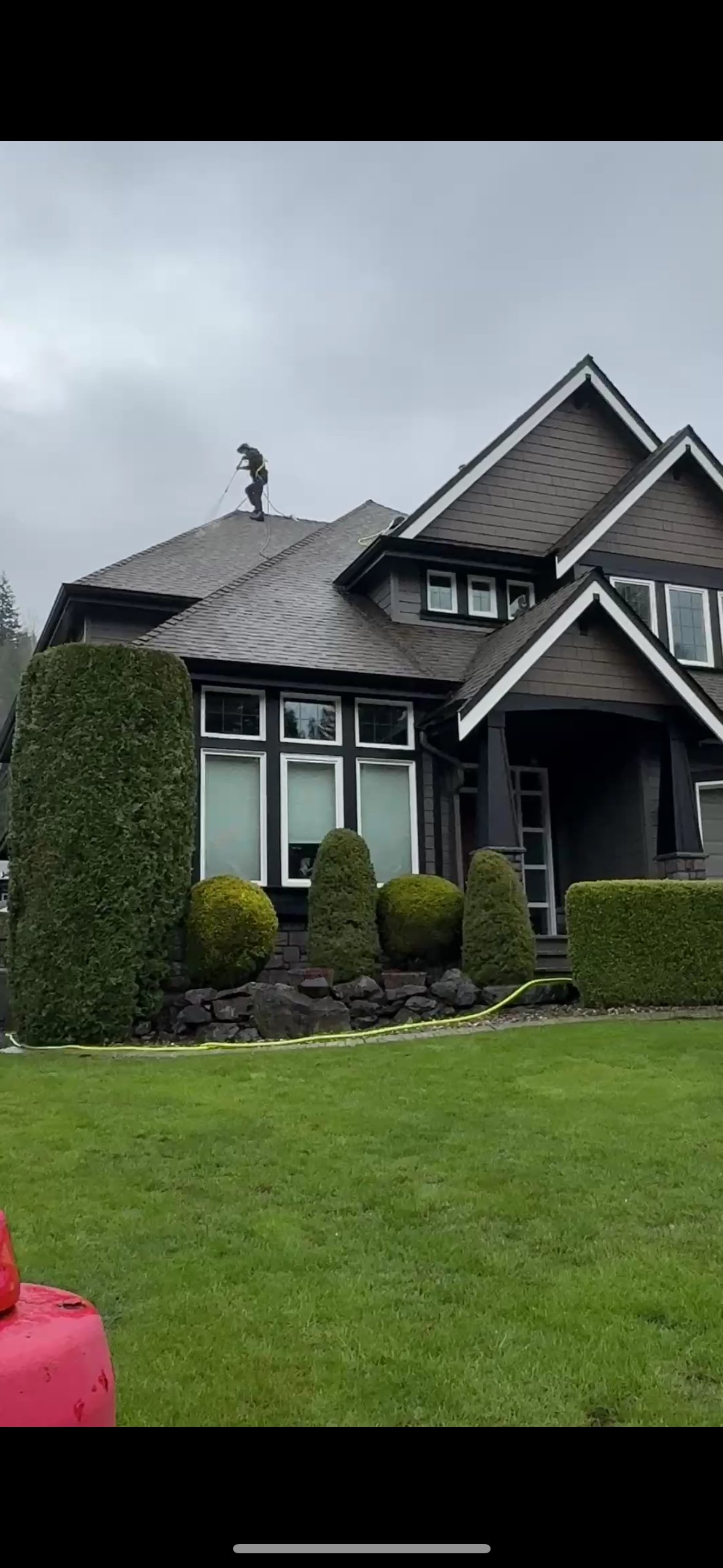 A person standing on the roof of a house, cleaning or inspecting it with a tool, under a cloudy sky.