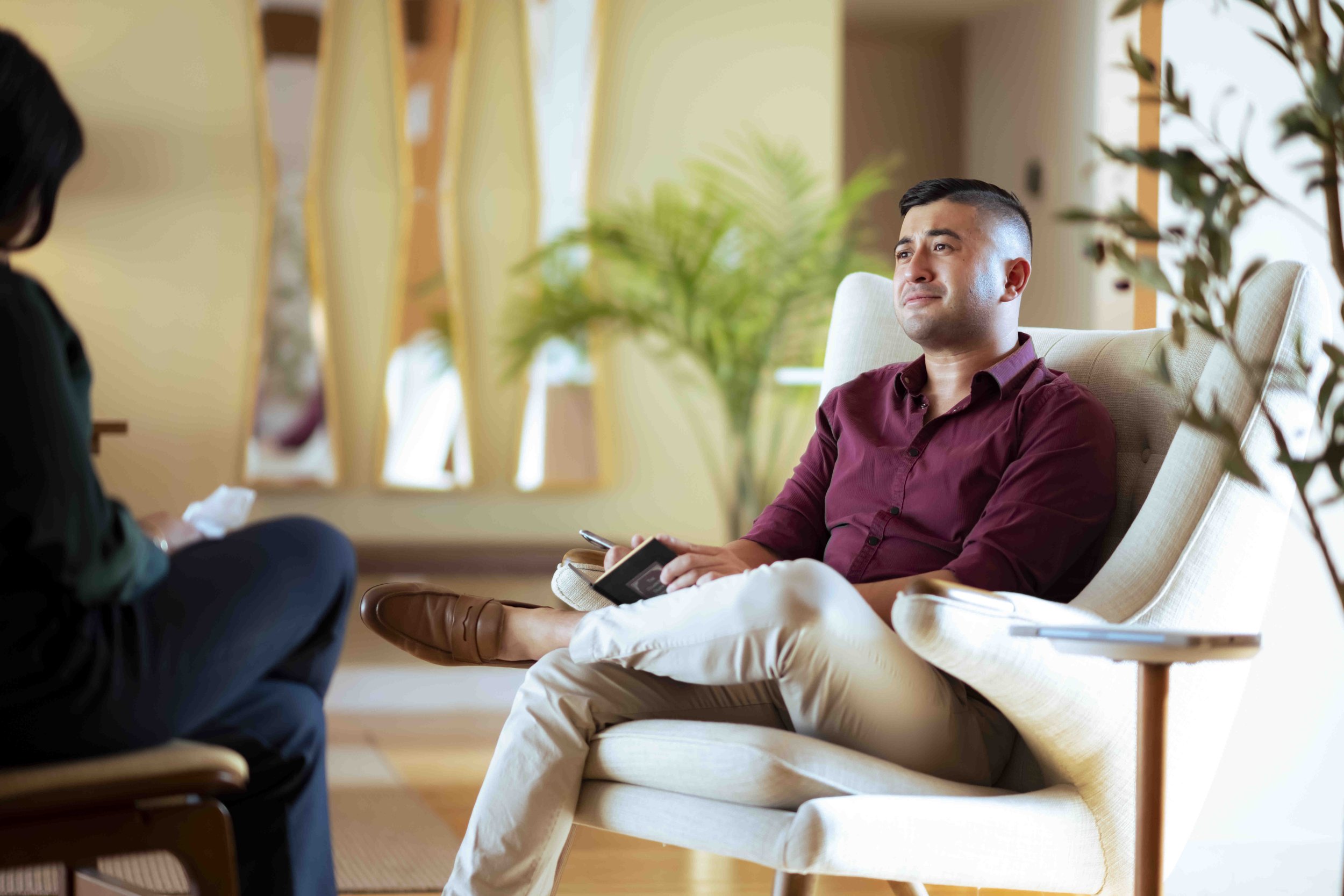 A man in a maroon shirt and beige pants sitting on a light-colored armchair in a bright room, holding a phone and a notepad, with a woman sitting opposite him, partially visible, in a conversation. There are plants and windows in the background.