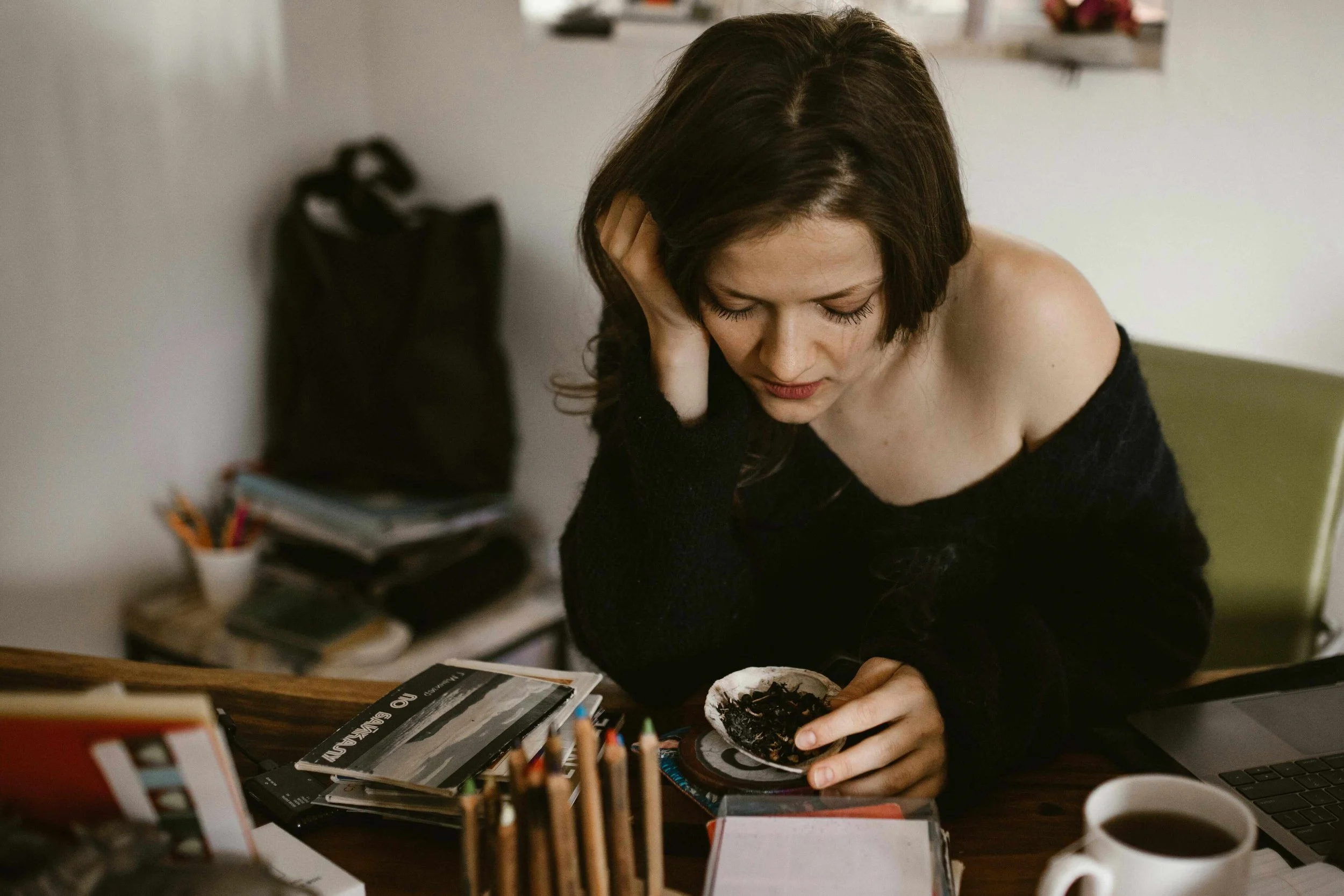 Woman with short brown hair, wearing a black off-the-shoulder sweater, sitting at a cluttered desk, holding a small bowl of food, with a laptop and a cup of coffee nearby.