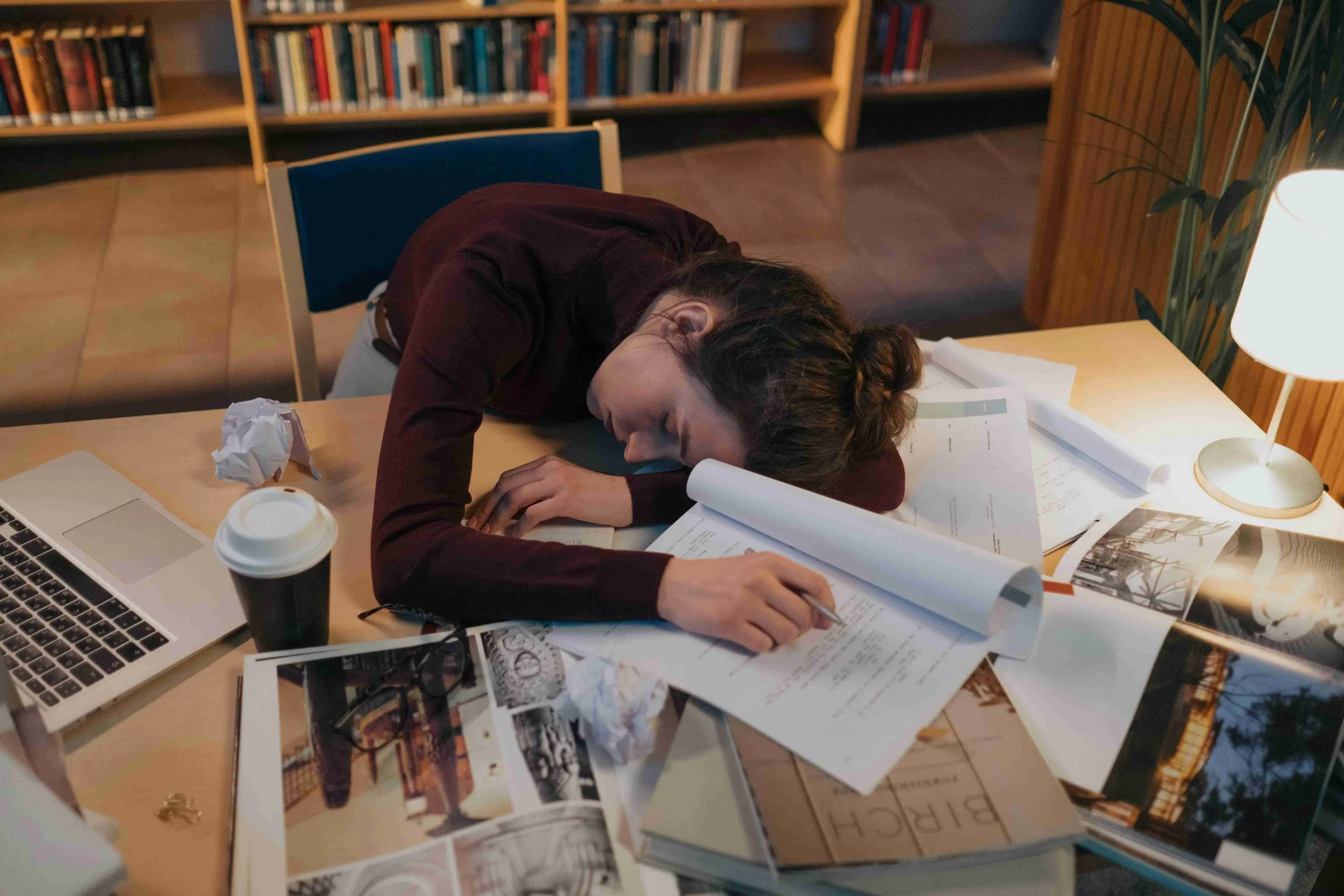 A woman is slumped over a cluttered desk with papers, a laptop, a coffee cup, glasses, and crumpled paper. She appears to be exhausted or asleep next to a lamp in a room with wooden shelves filled with books.