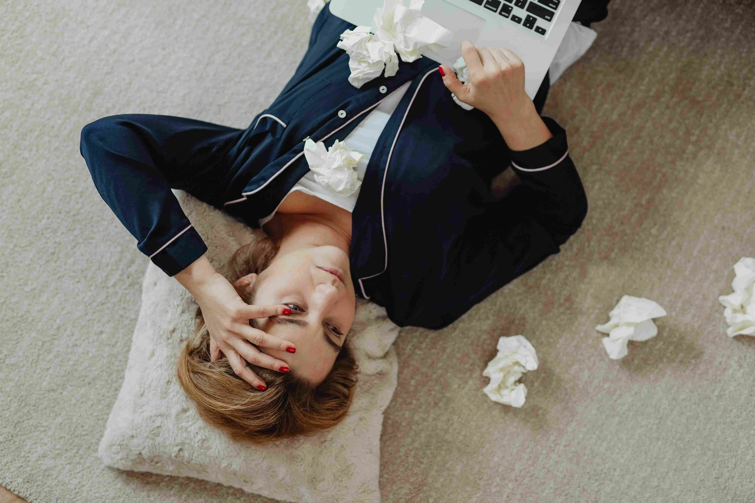 A woman lying on her back on a beige carpeted floor, surrounded by crumpled tissues, wearing navy blue pajamas with white piping, holding a laptop, with her left hand on her forehead and her eyes slightly squinted, expressing fatigue or sickness.
