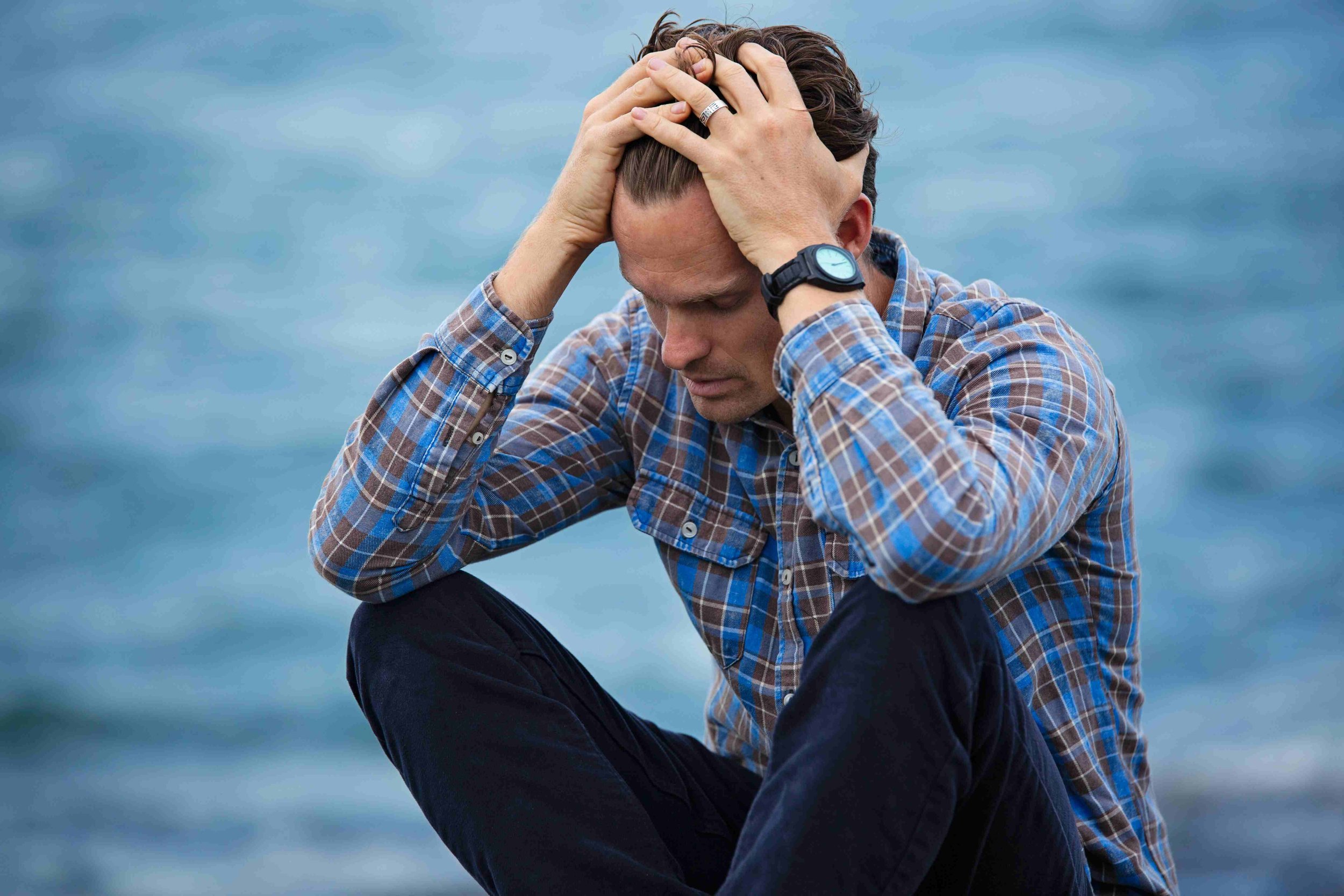 A man sitting by the water with a distressed expression, holding his head with both hands, wearing a plaid shirt and a watch.