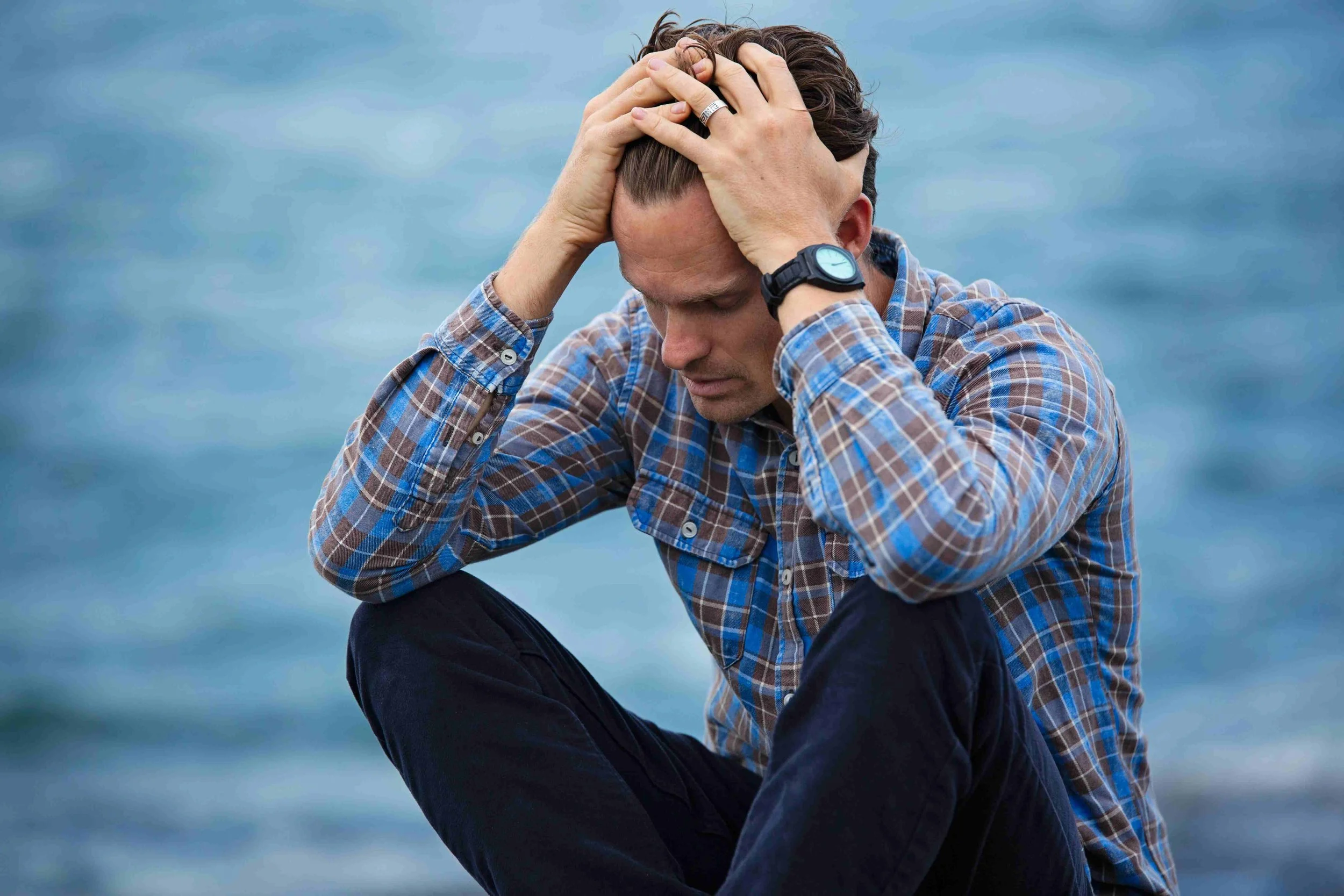 A man sitting by the water with a distressed expression, holding his head with both hands, wearing a plaid shirt and a watch.