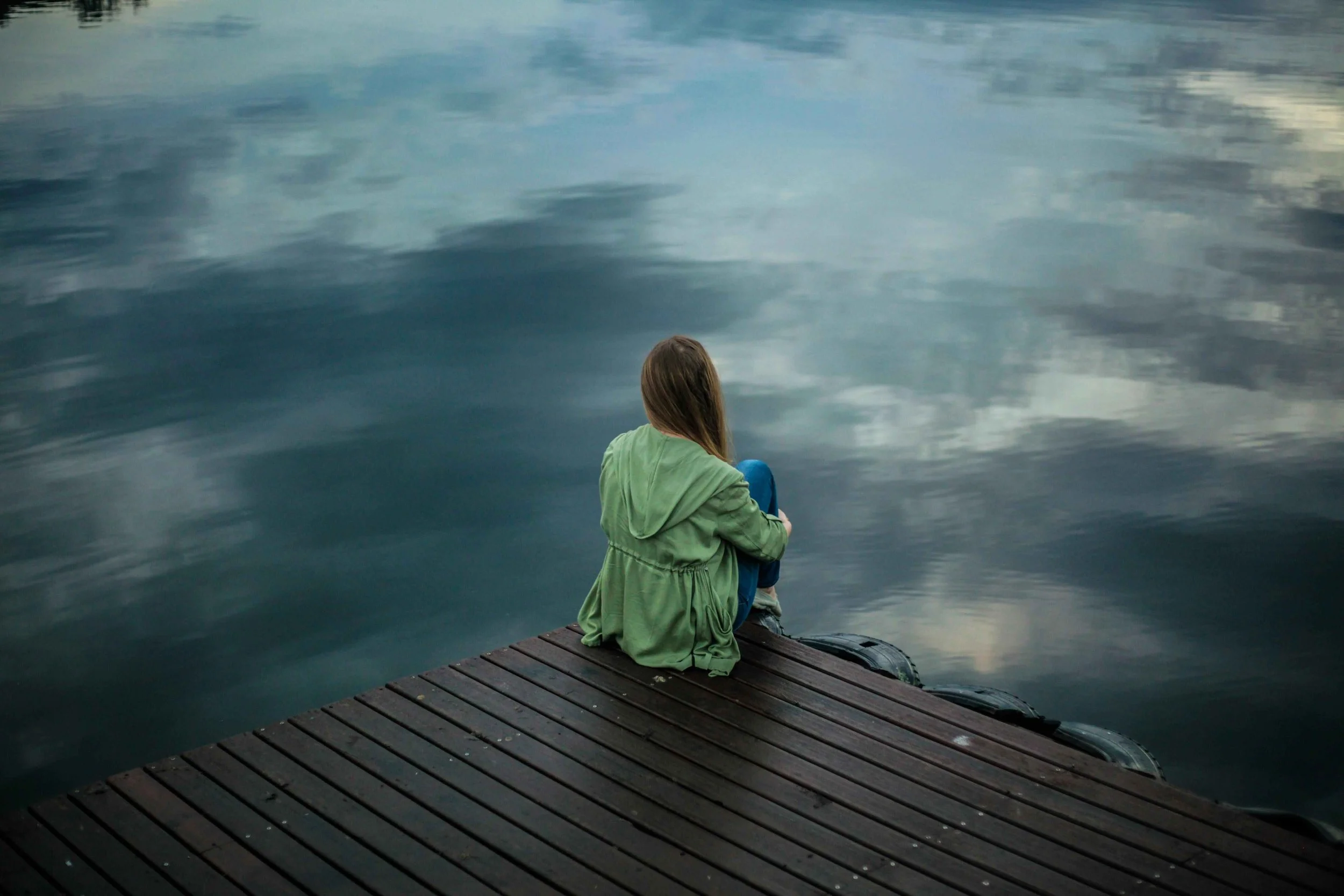 A woman with long hair sitting alone on a wooden dock, looking at calm water with reflections of the sky.