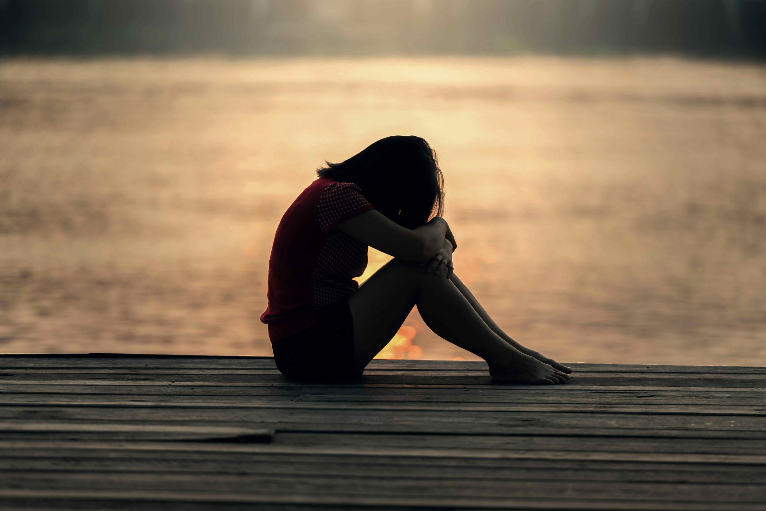 Silhouette of a woman sitting on a wooden dock by the water, hugging her knees with her head bowed during sunset.