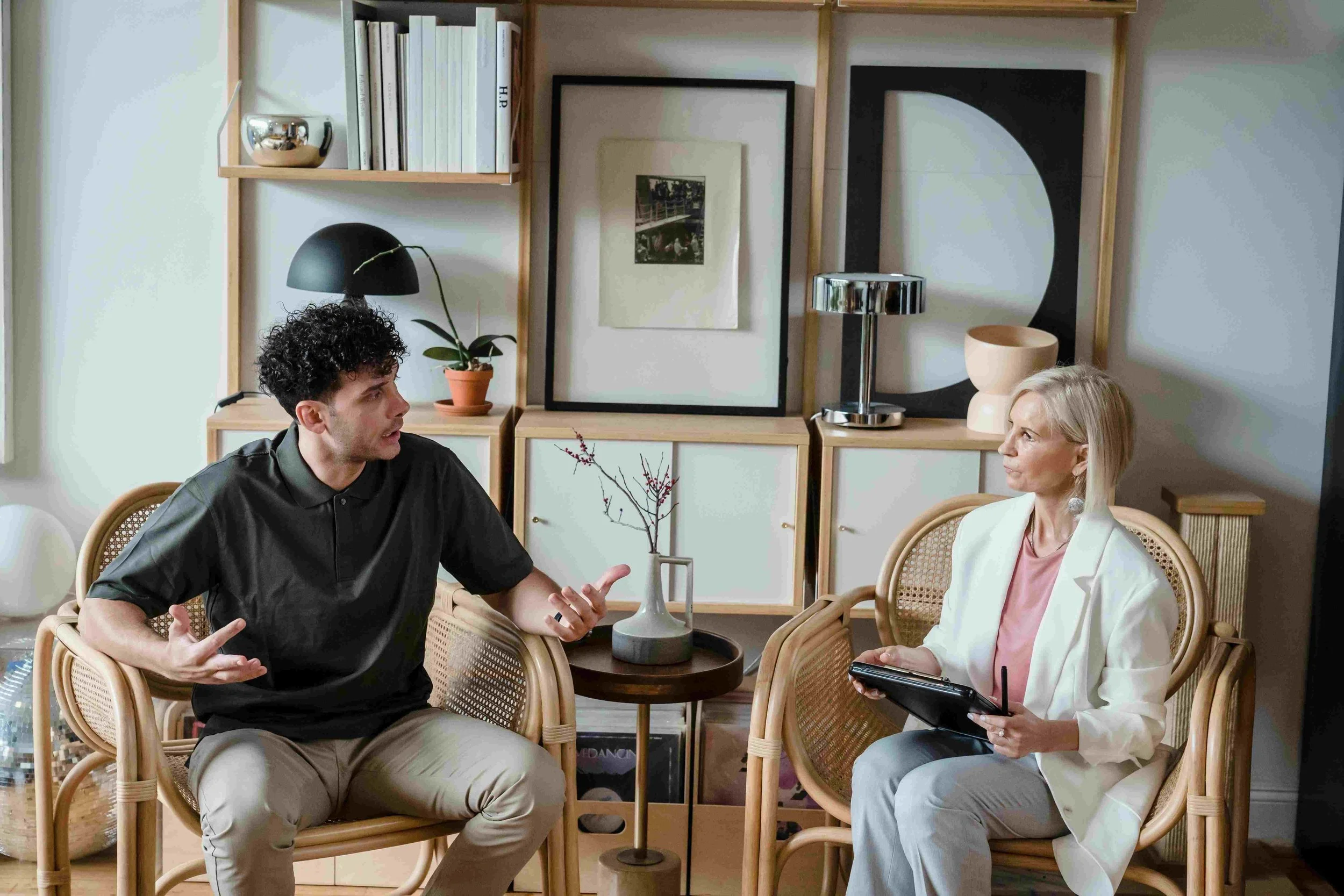 A man and a woman seated and having a conversation in a living room with modern decor, including framed artwork, plants, and wooden shelving.