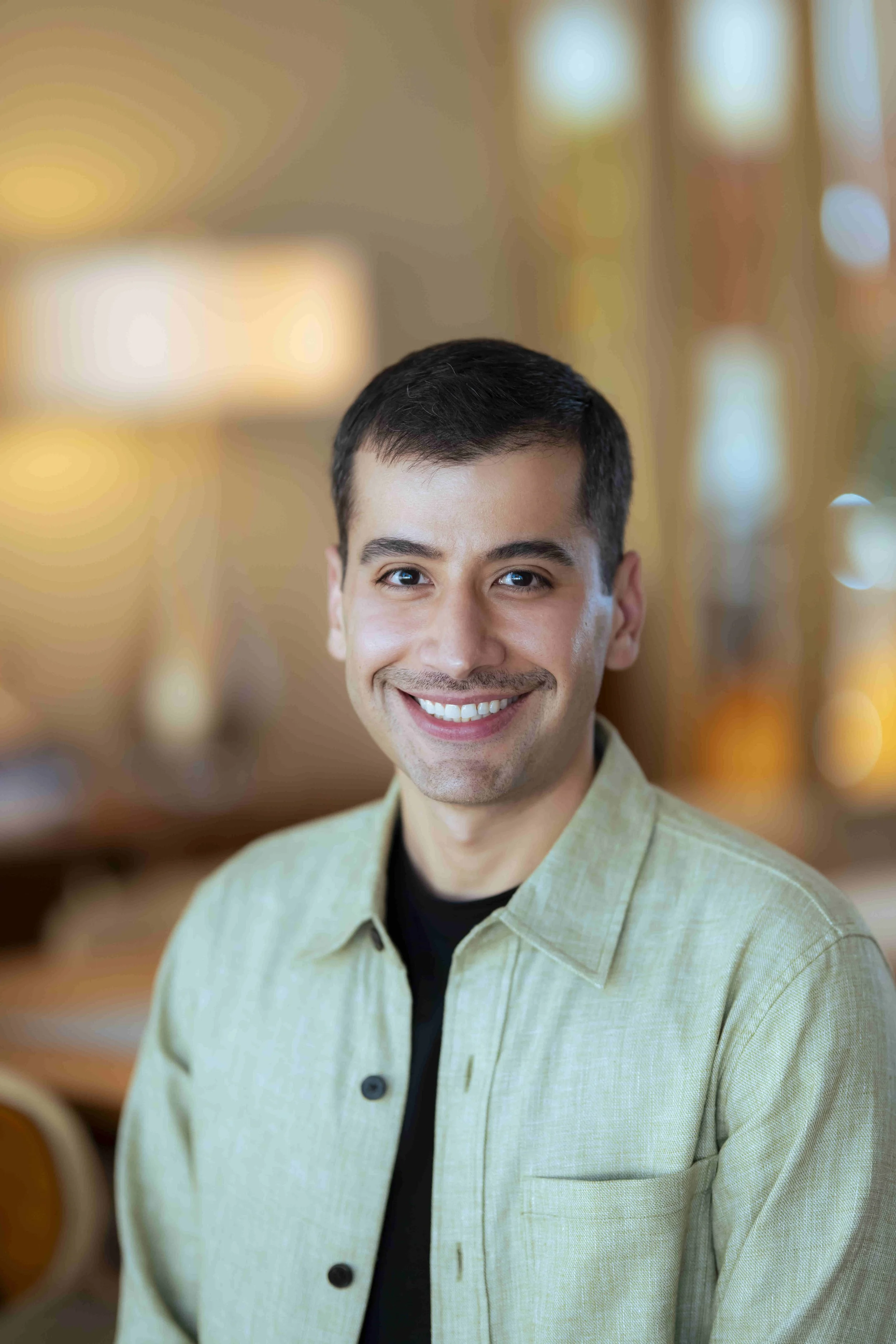 A smiling man with short dark hair, wearing a light green button-up shirt over a black t-shirt, in a warmly lit indoor setting.