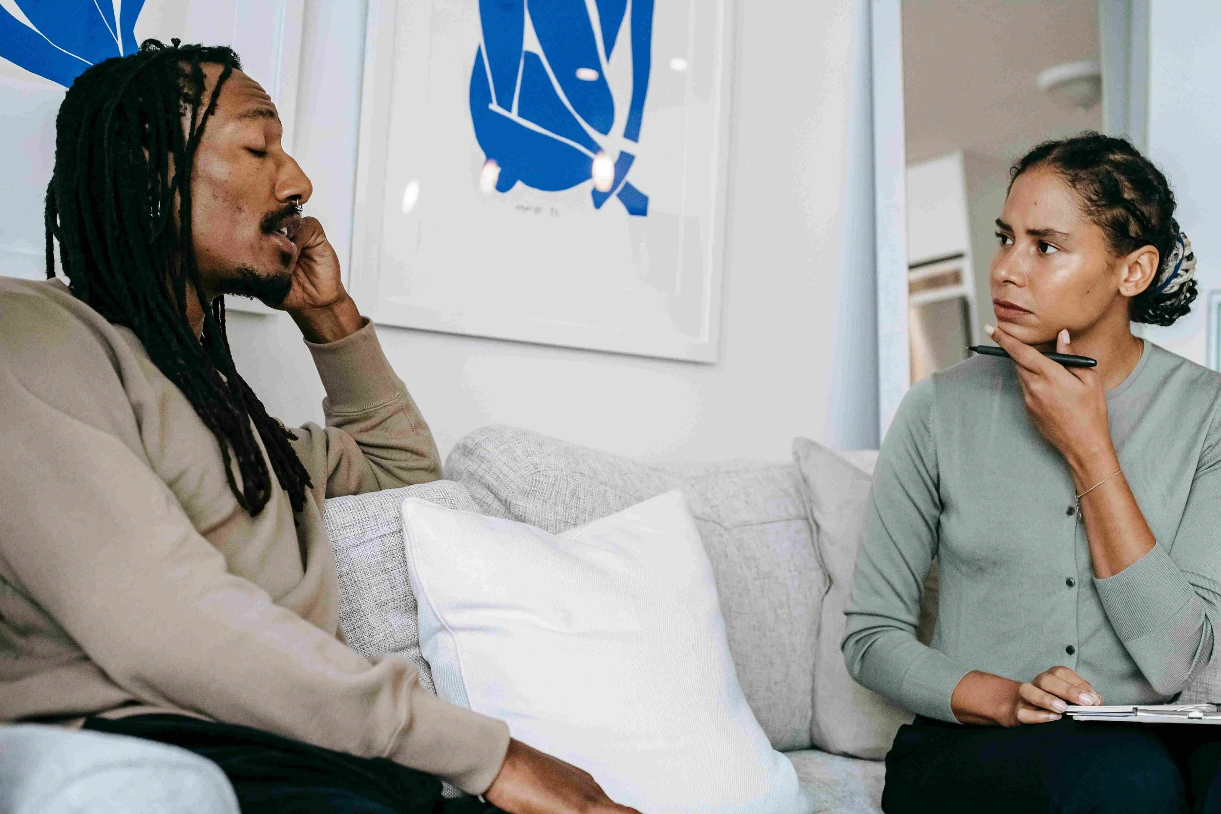 A man and a woman having a serious conversation in a living room. The man, with dreadlocks, is sitting on a couch with his hand on his face. The woman, with curly hair, is holding a pen and a notepad, and looks concerned or questioning.