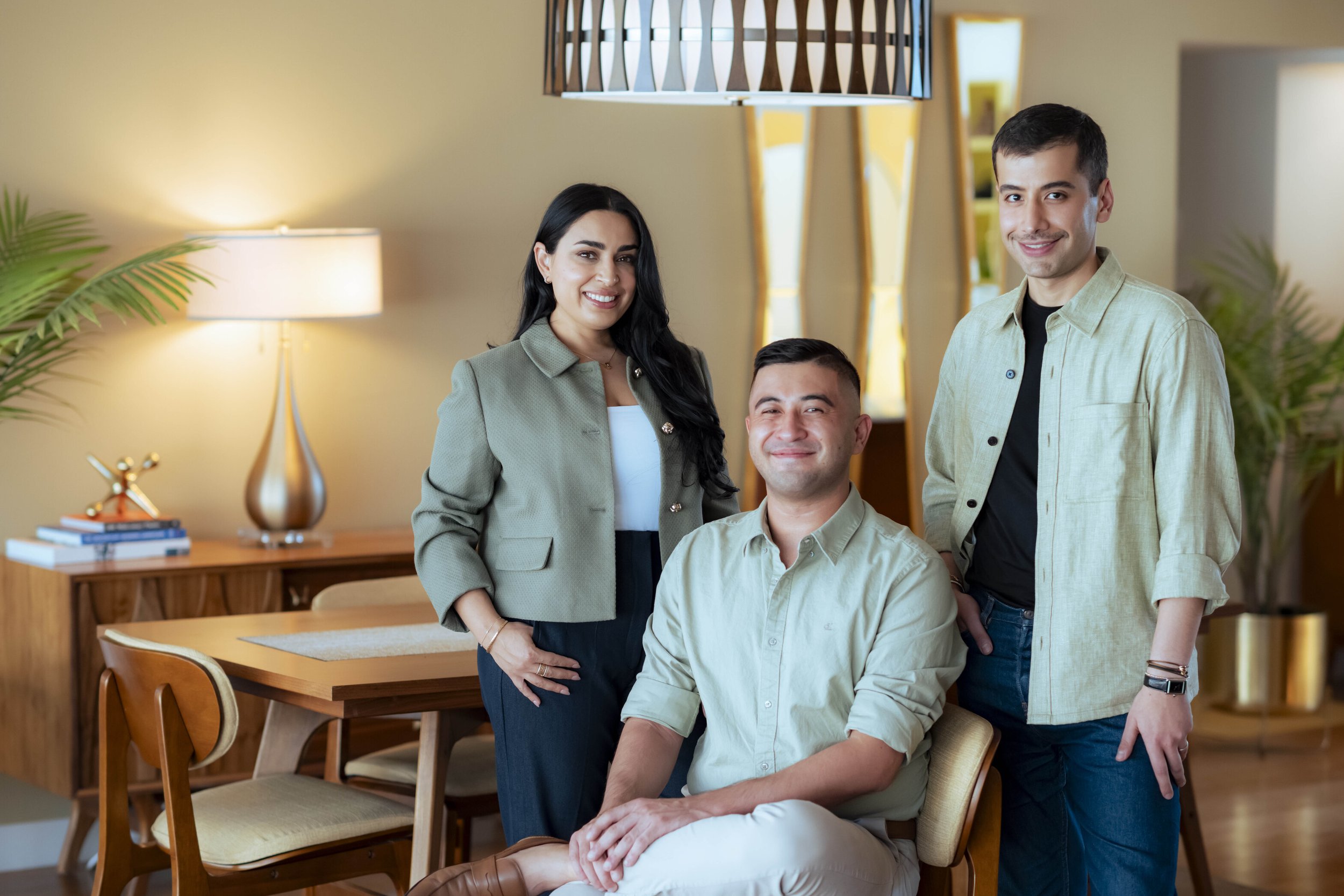 Three therapists, two men and one woman, smiling and posing in a warmly lit room with a modern decor. One man is seated in a chair, while the others stand behind him. The room has a wooden dining table, chairs, a lamp, plants, and mirrors.