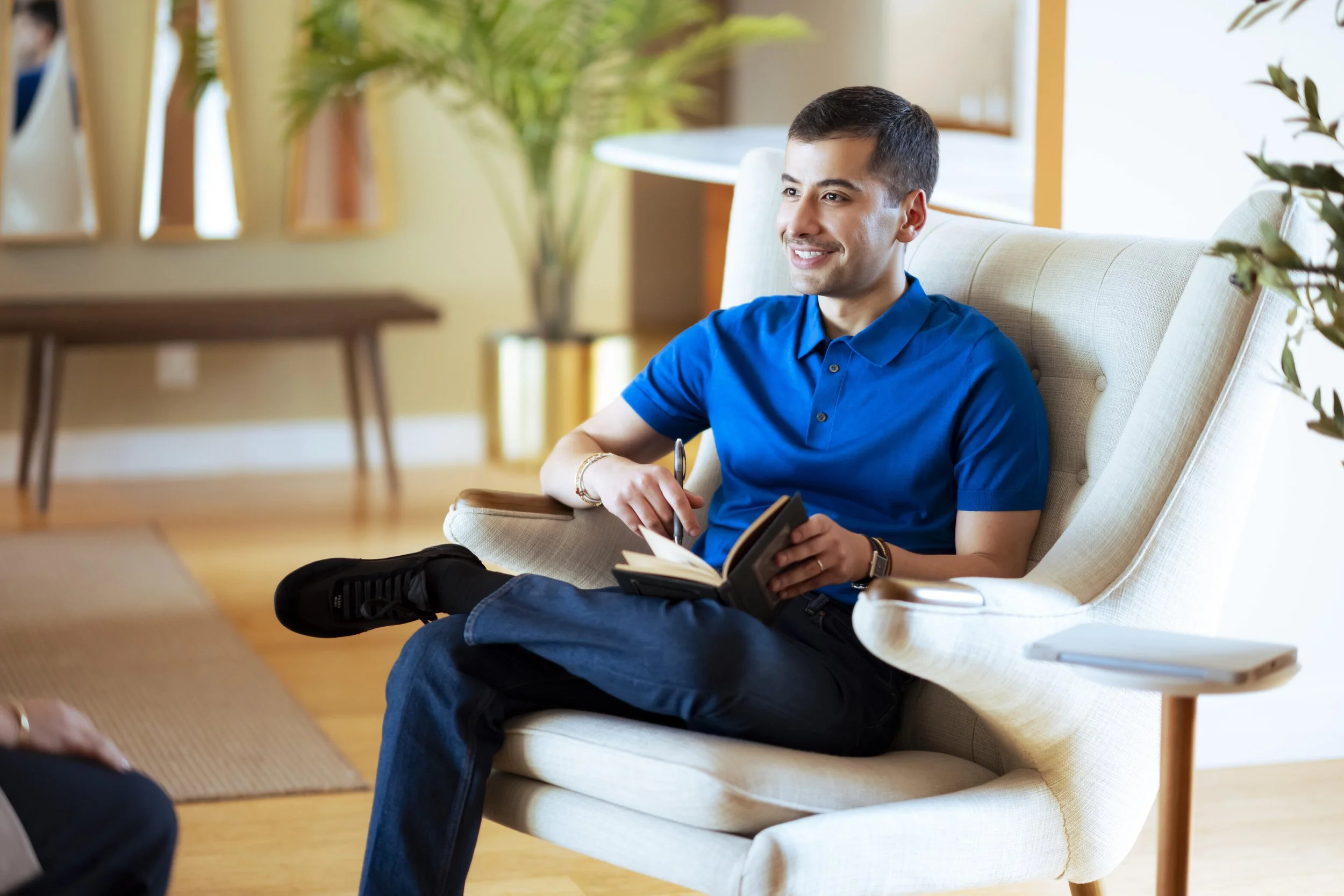 A smiling male therapist in a blue polo shirt sitting on a beige couch, holding a small notebook and a pen, with his legs crossed in a well-lit, modern living room with plants and wooden furniture.