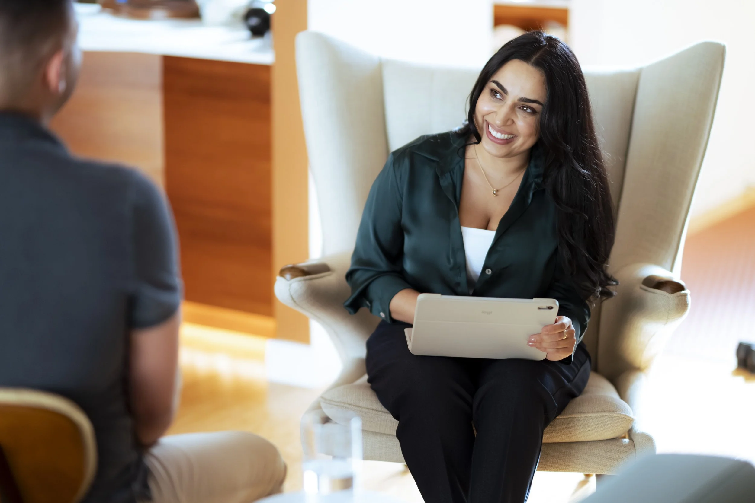 A female therapist during a conversation while holding a tablet, sitting in a high-backed chair, with another person partially visible in the foreground.