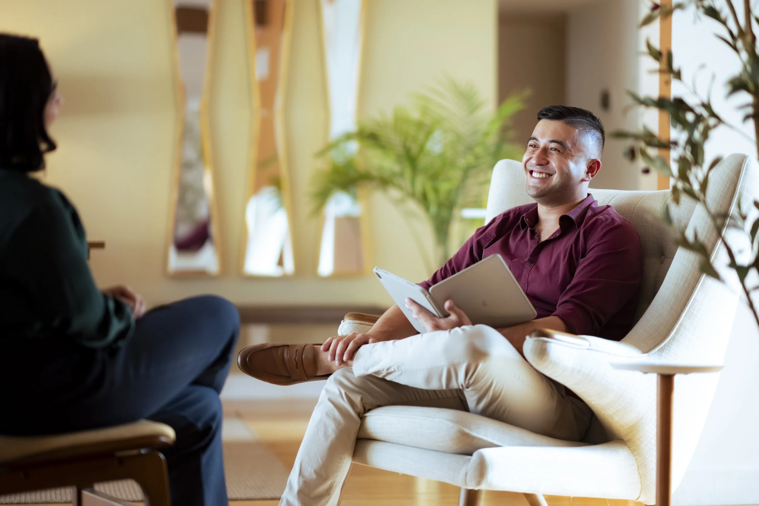 Man smiling during a therapy or counseling session with a woman in a cozy, well-lit room with plants and decorative mirrors.