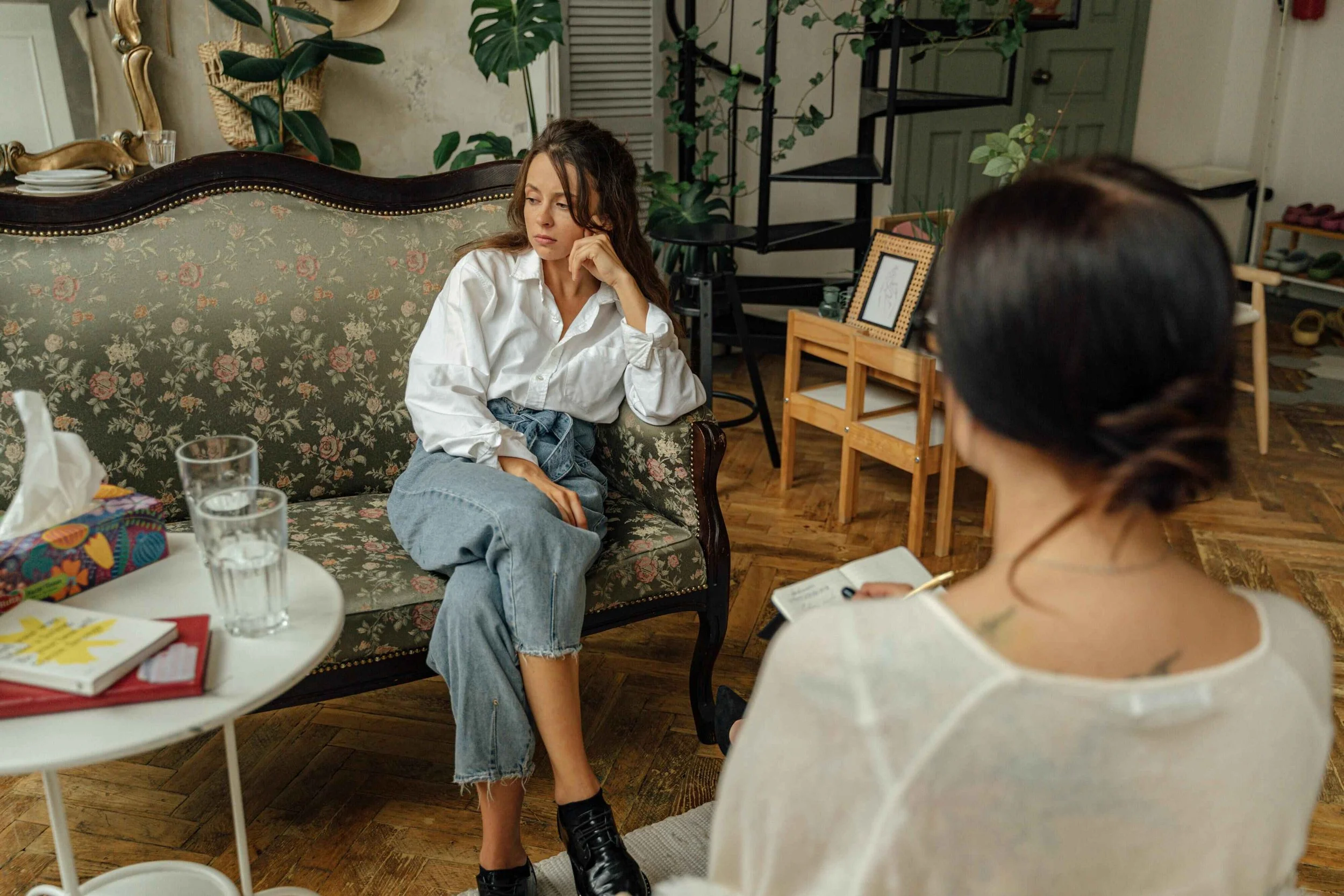 A woman sitting on a floral upholstered sofa during a therapy or counseling session, listening attentively to her therapist who is taking notes, in a cozy room with plants and wooden furniture.