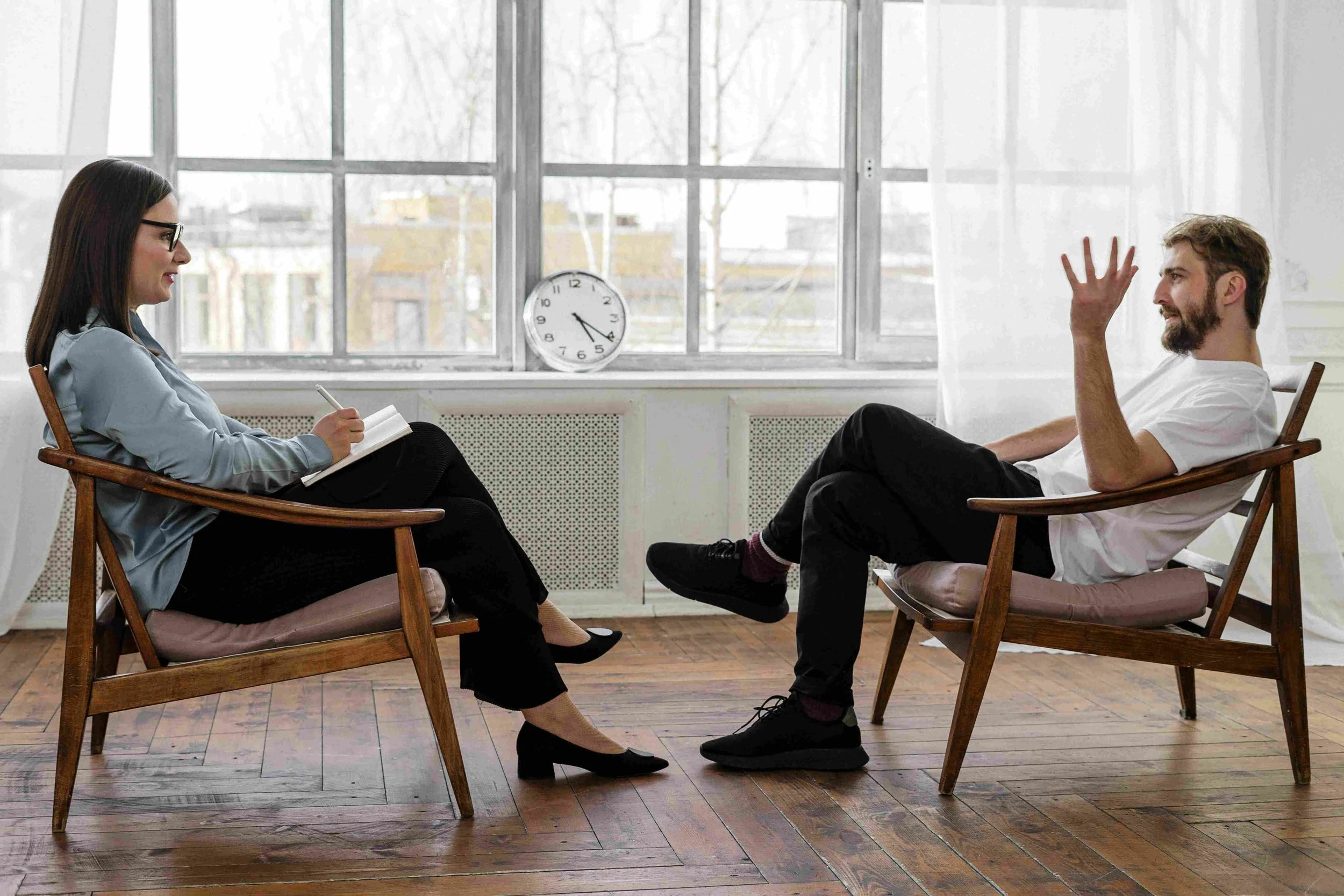 Therapist sitting on a wooden chair with a notebook, talking to a male patient in a white T-shirt, sitting on a similar chair, inside a room with large windows, a clock on the windowsill, and wooden flooring.