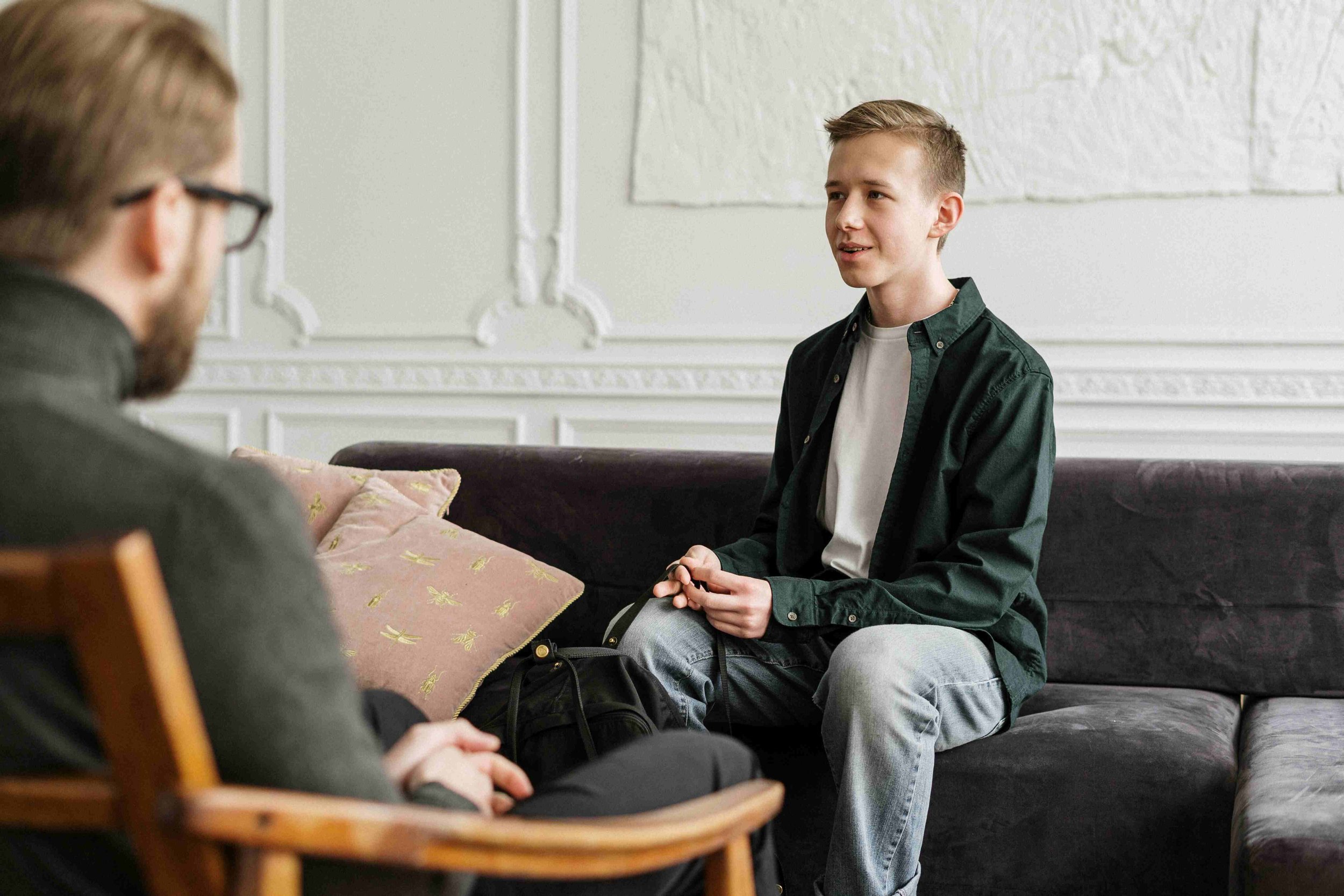 Two males having a conversation in a room with white walls, sitting on a dark-colored sofa and a wooden chair. One is an older male with glasses, and the other is a younger male with short blonde hair, wearing a green shirt over a white t-shirt.