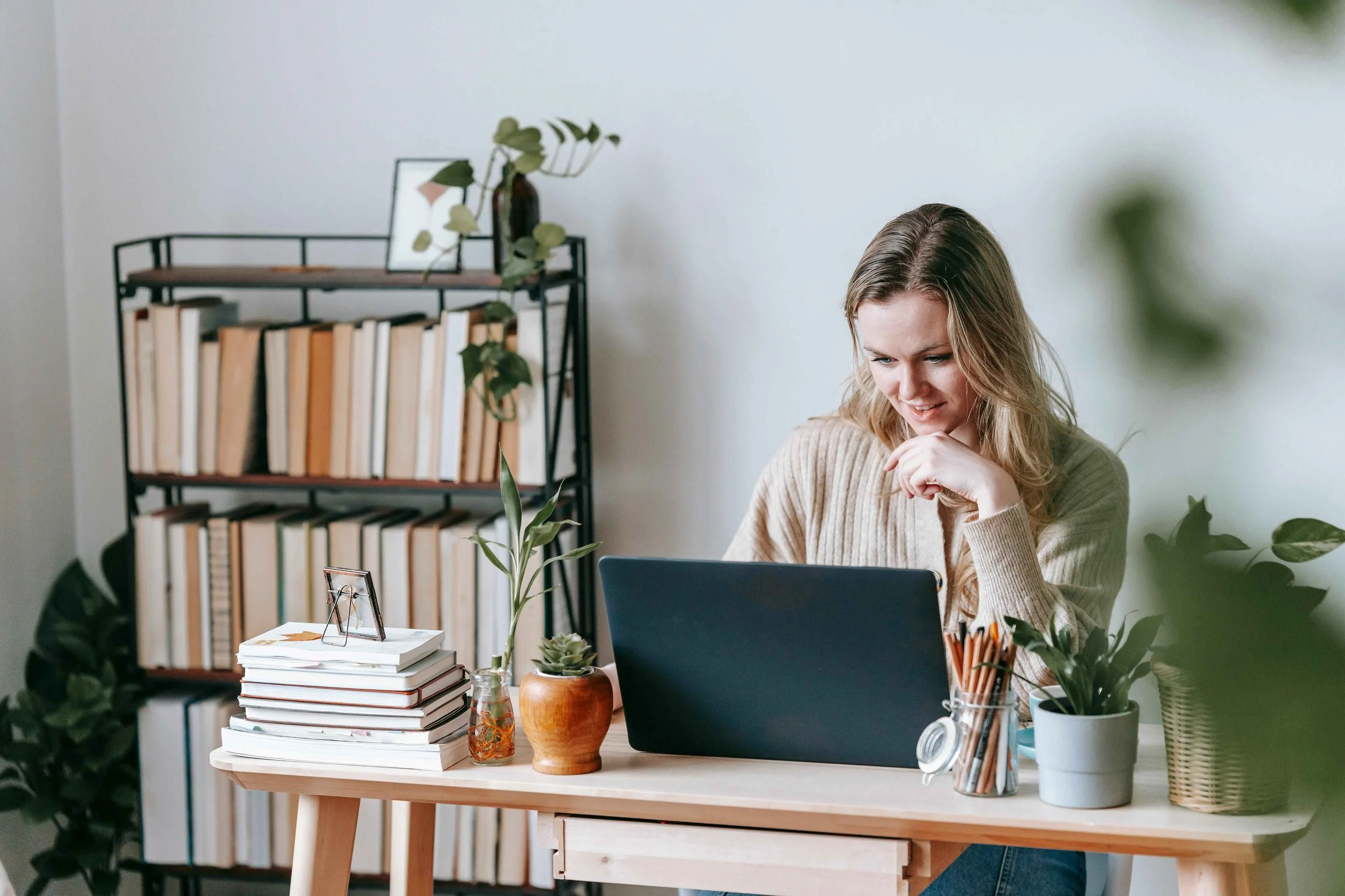 Woman working at a desk with plants, notebooks, and a laptop in a home office setting.