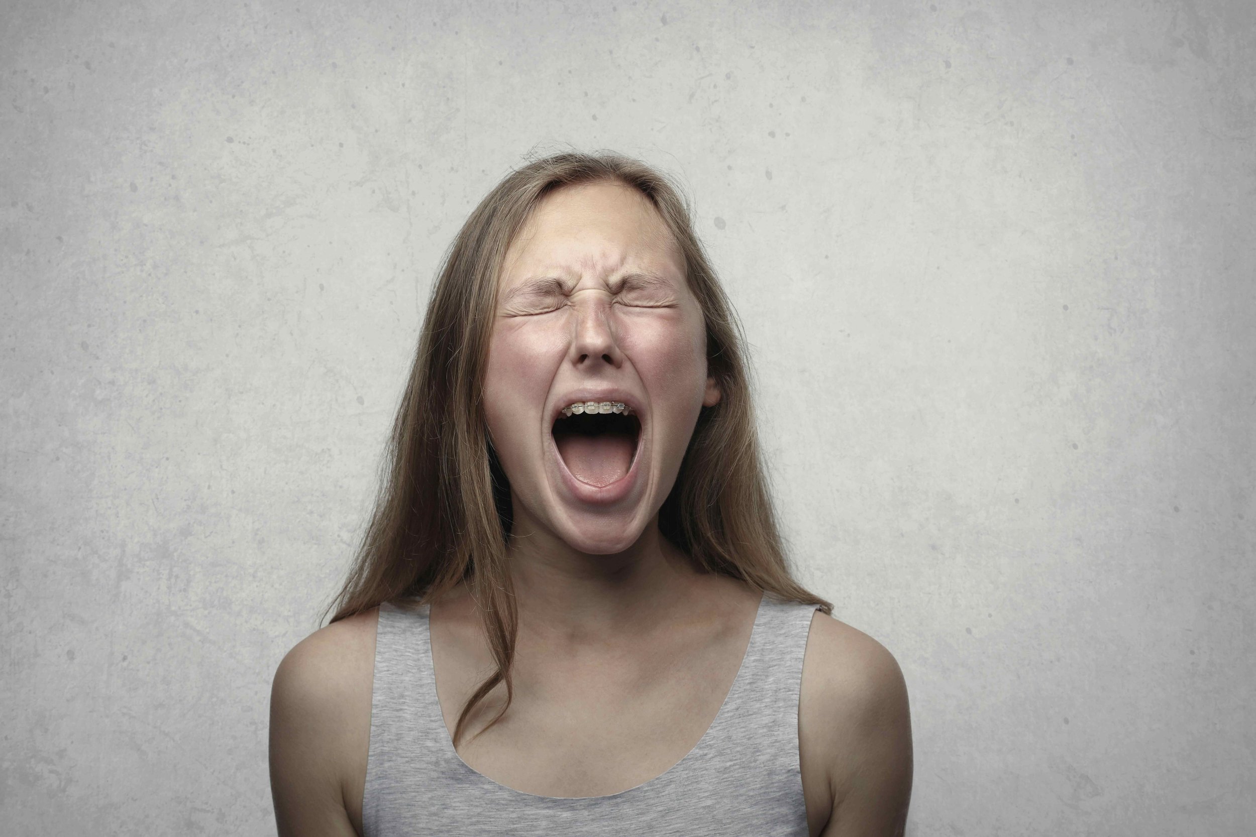 A young woman with long brown hair wearing a gray tank top is shouting or yawning with her eyes closed against a plain, light gray background.