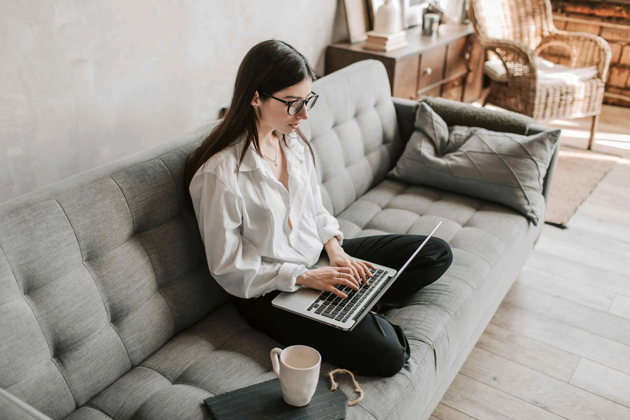 A woman sitting cross-legged on a grey sofa working on a laptop, with a coffee mug and a small black tray nearby, in a cozy living room with wooden floors and wicker chairs.