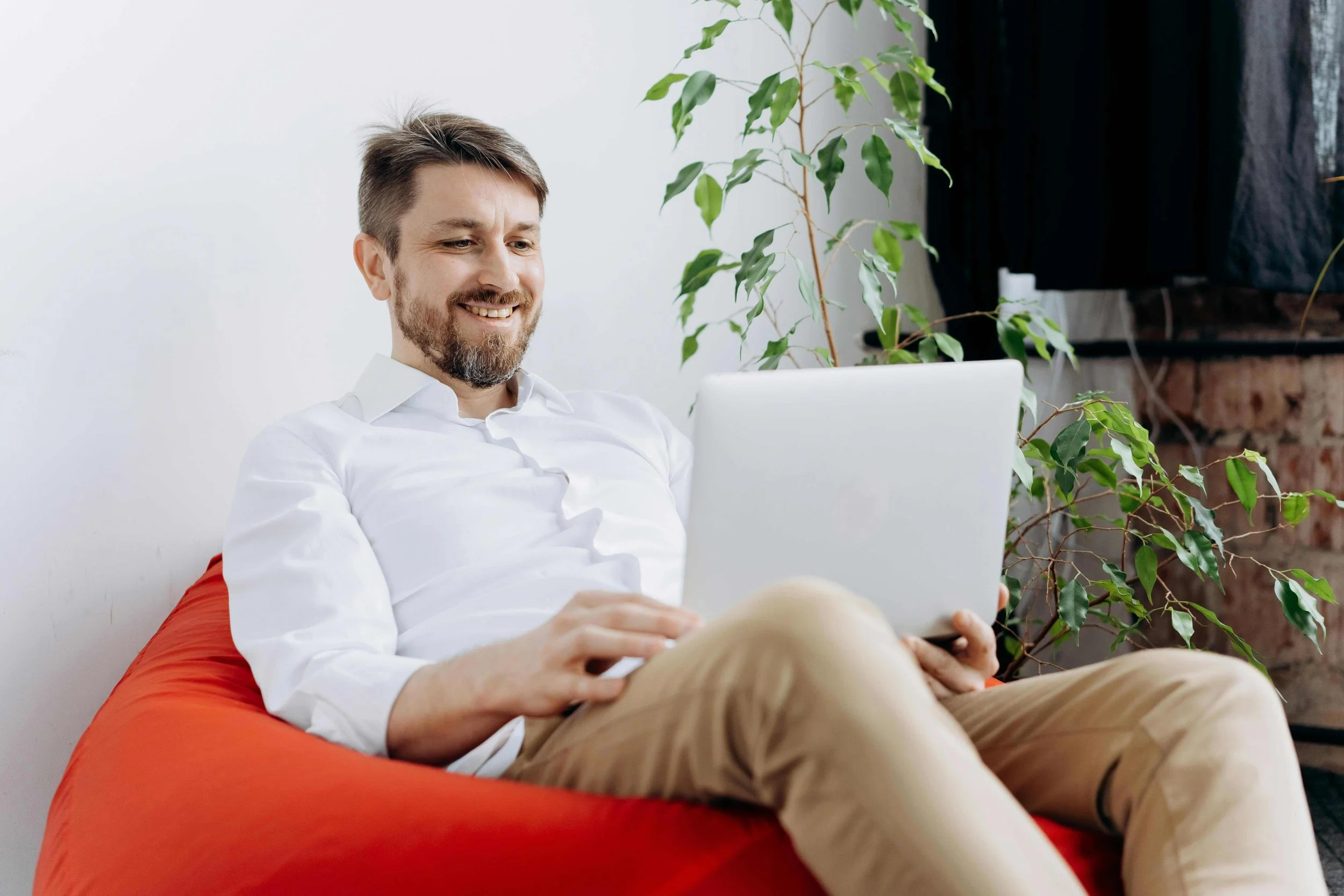 A man with a beard and short hair is sitting on a red cushion, smiling and using a silver laptop. There is a green plant nearby and a white wall behind him.
