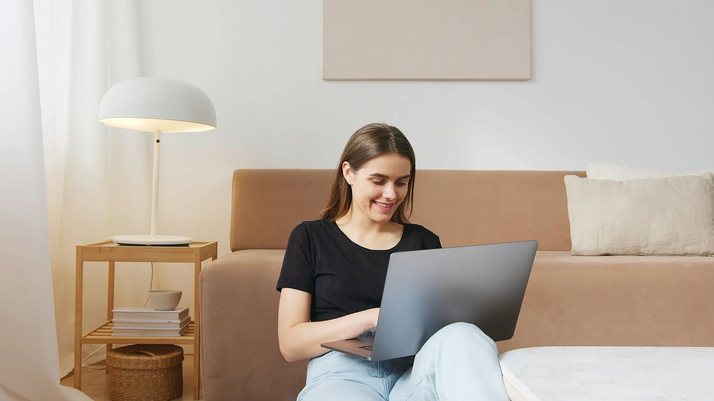 A woman with long brown hair smiling while using a laptop in a cozy, minimalist living room with a beige sofa, a wooden side table, a white lamp, and light-colored pillows.