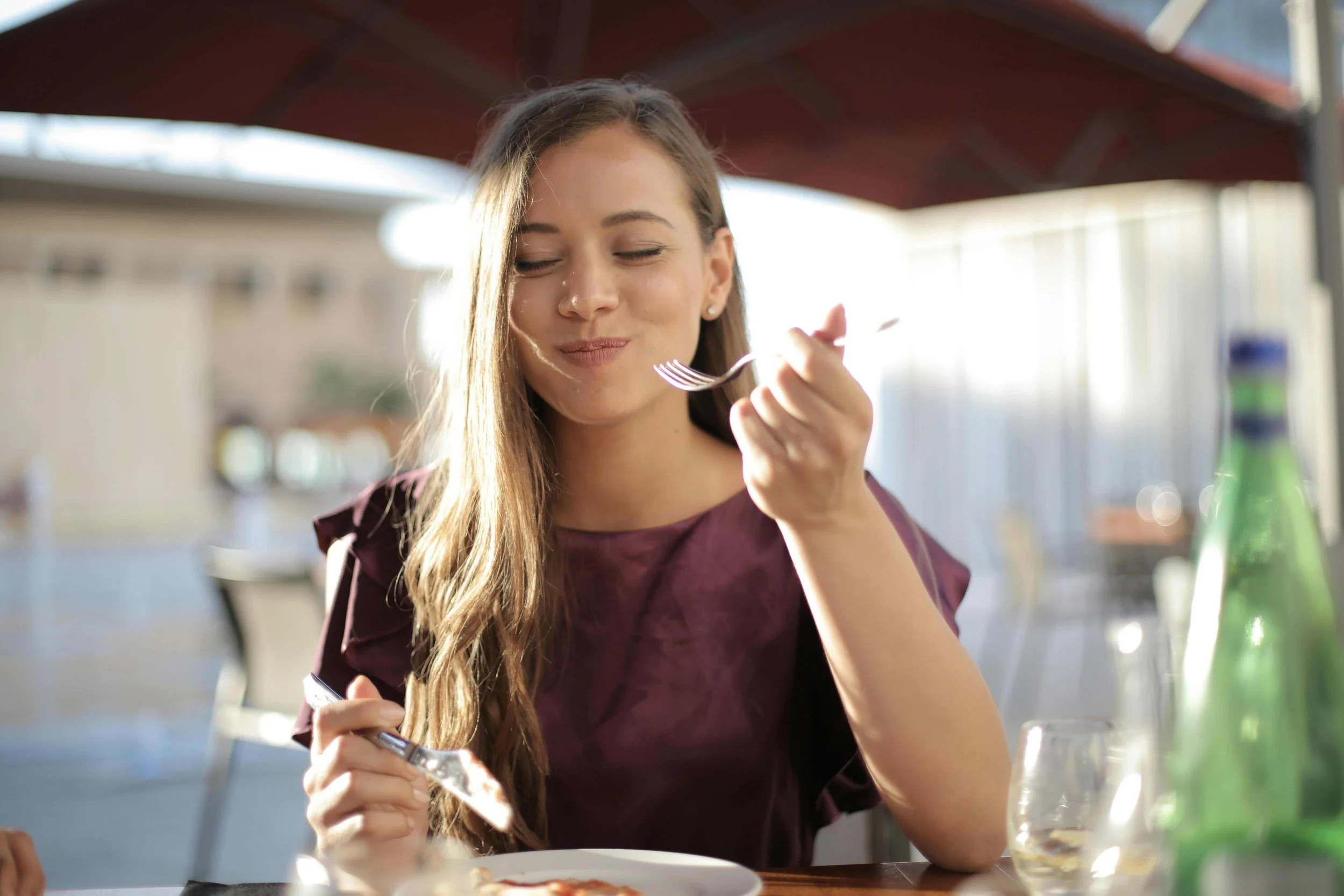 A young woman with long hair enjoying a meal at an outdoor restaurant, smiling with her eyes closed, holding a fork in her right hand.