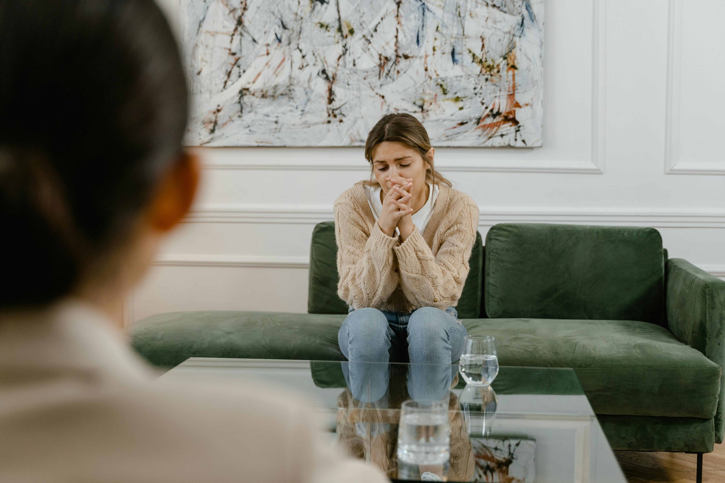 A young woman with brown hair sitting on a green couch with her hands clasped in front of her face, appearing distressed or upset, during a discussion or therapy session in a room with white walls and an abstract painting behind her. There are two glasses of water on a glass table in front of her, and another person is partially visible in the foreground, out of focus.