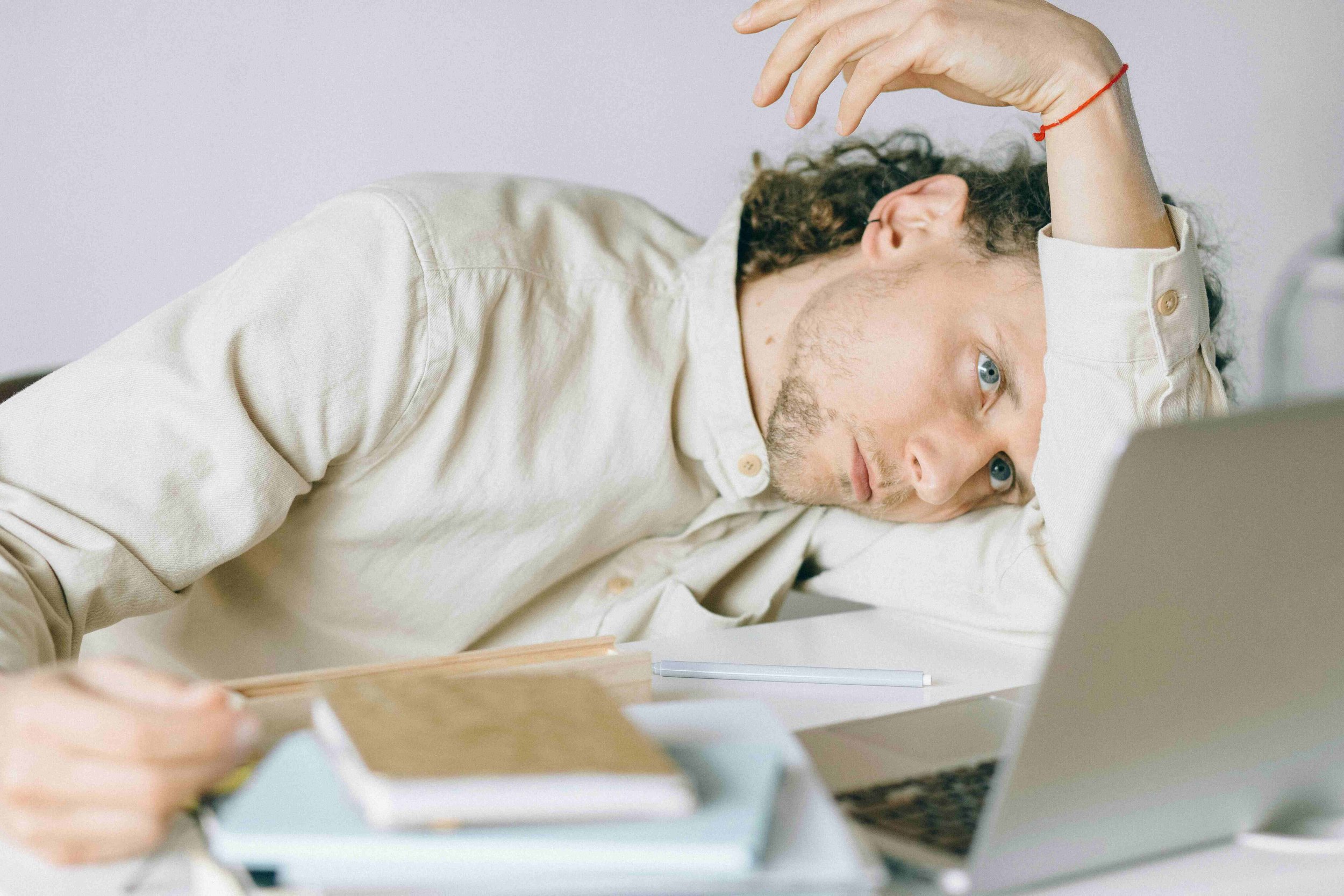 A man with curly hair and a beard is lying on a desk with his head resting on his arm, looking tired or stressed. He is wearing a white shirt and is surrounded by a laptop, notebooks, and a pen.