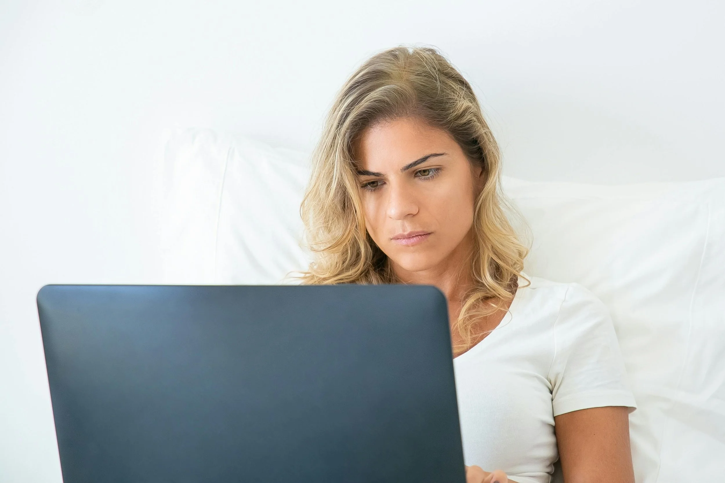 A young woman with blonde hair sitting in bed, looking at a laptop with a serious expression.