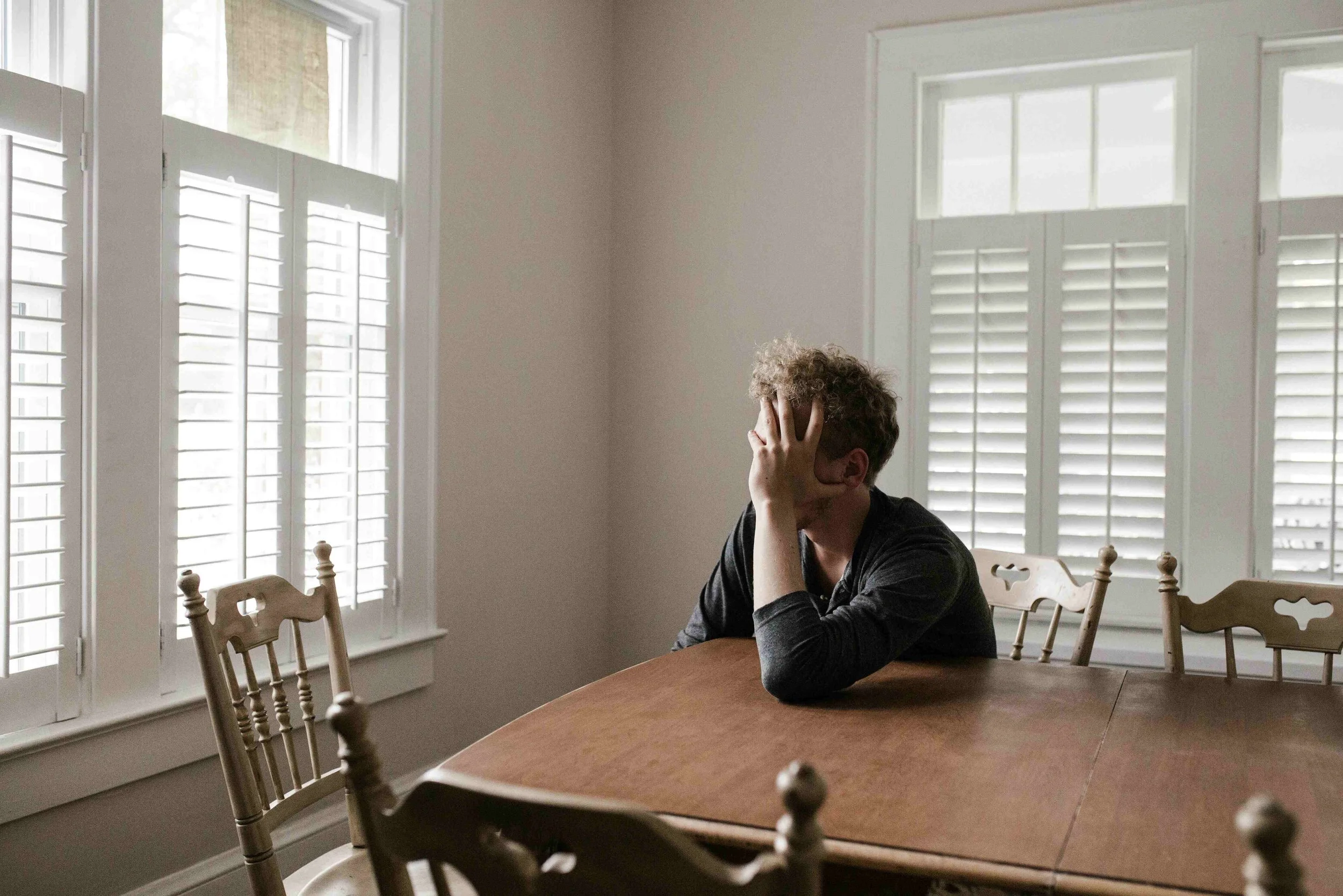 A man sitting at a dining table with his head in his hand, appearing distressed or frustrated, in a room with white walls and large windows with shutters.
