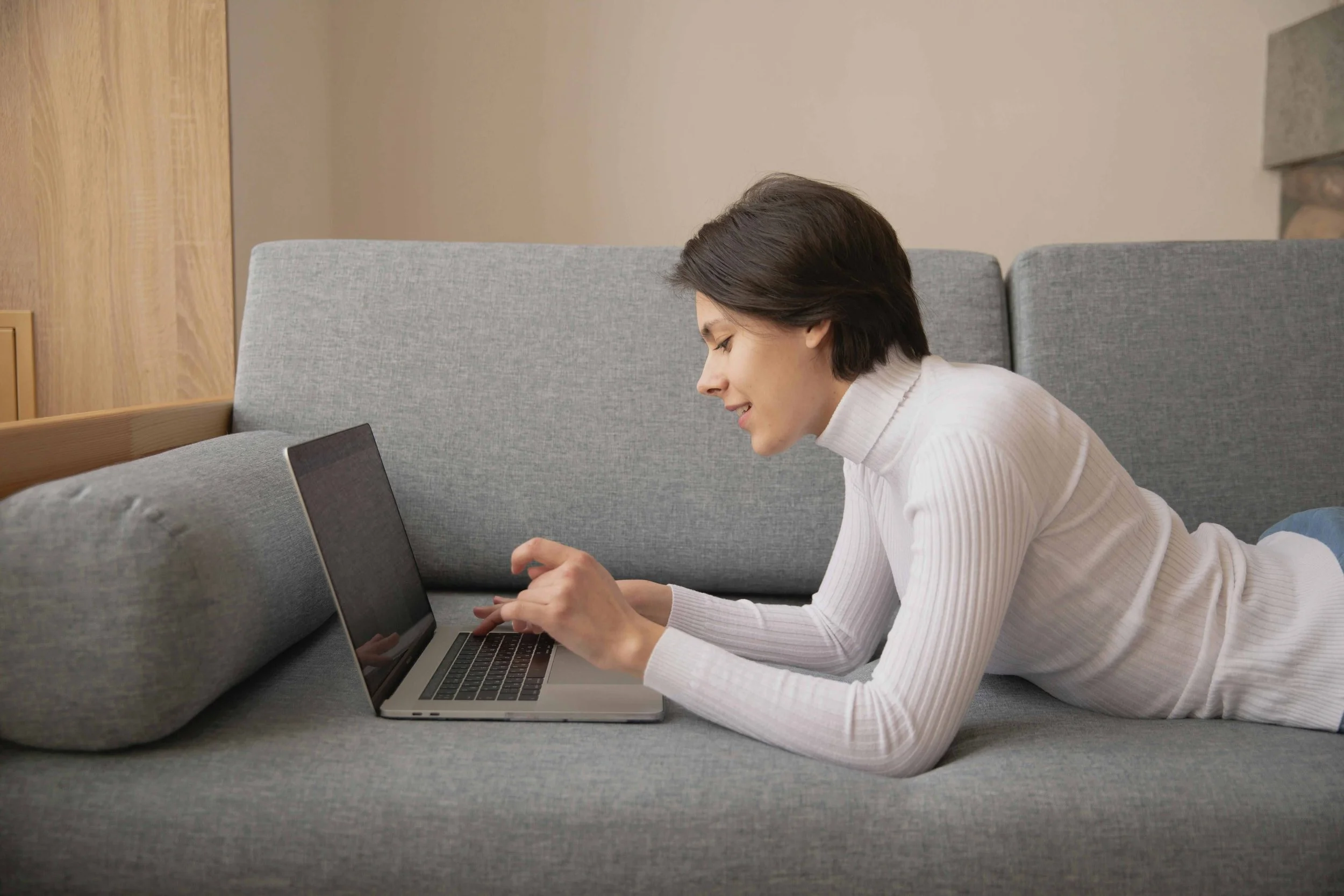 Woman lying on her stomach on a gray couch, using a laptop.