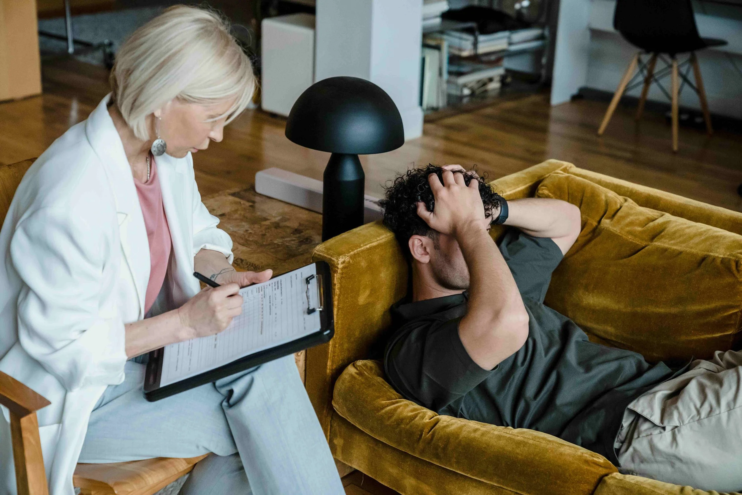 A woman in a white blazer sitting on a chair taking notes on a clipboard while a man in a black shirt lies on a mustard-colored sofa holding his head in distress. The setting appears to be a therapy or counseling session in a living room.