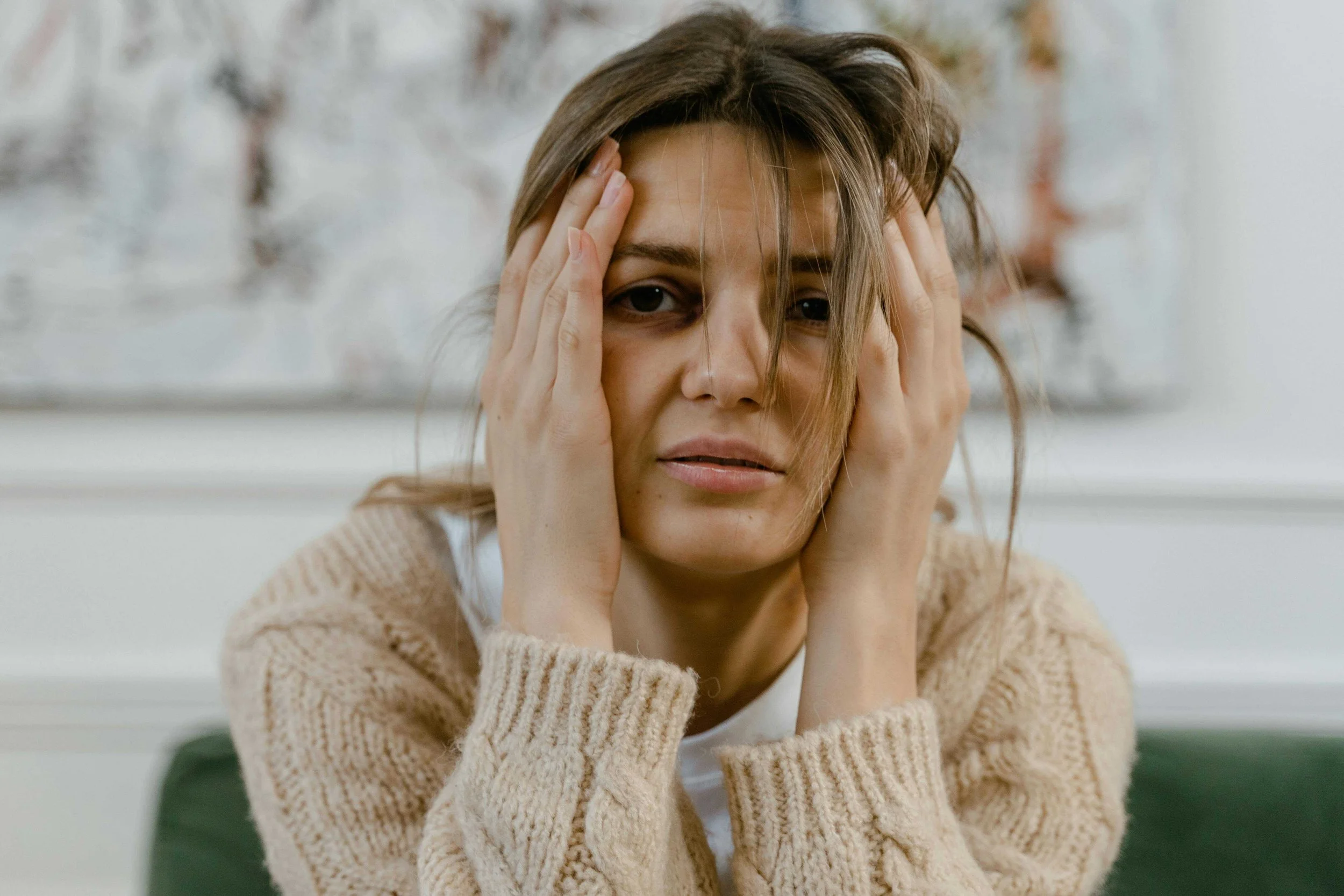 A woman with brown hair, wearing a beige sweater, holding her head with both hands, appearing stressed or overwhelmed.